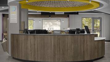 A modern hotel reception desk with a wooden finish, a yellow accent wall, and a sign indicating it is a Hilton Honors hotel.