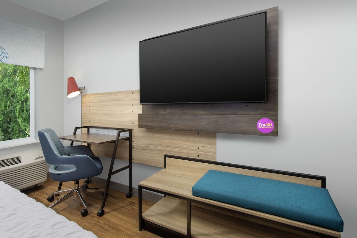 Un dormitorio moderno con cama, escritorio, silla, televisión y ventana con vistas a zonas verdes.