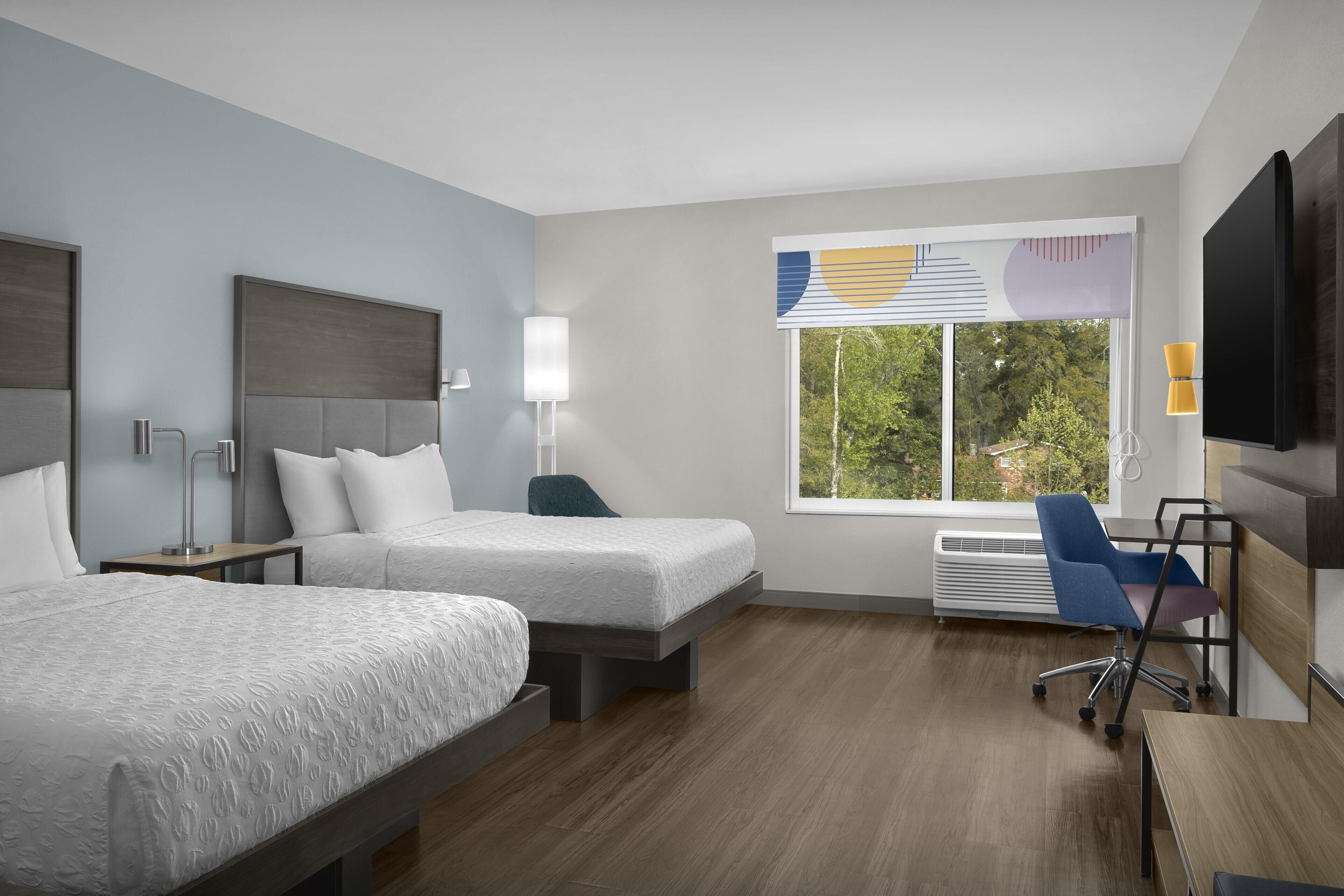 A hotel room with two beds, a desk, a chair, a TV, and a window with a view of trees.