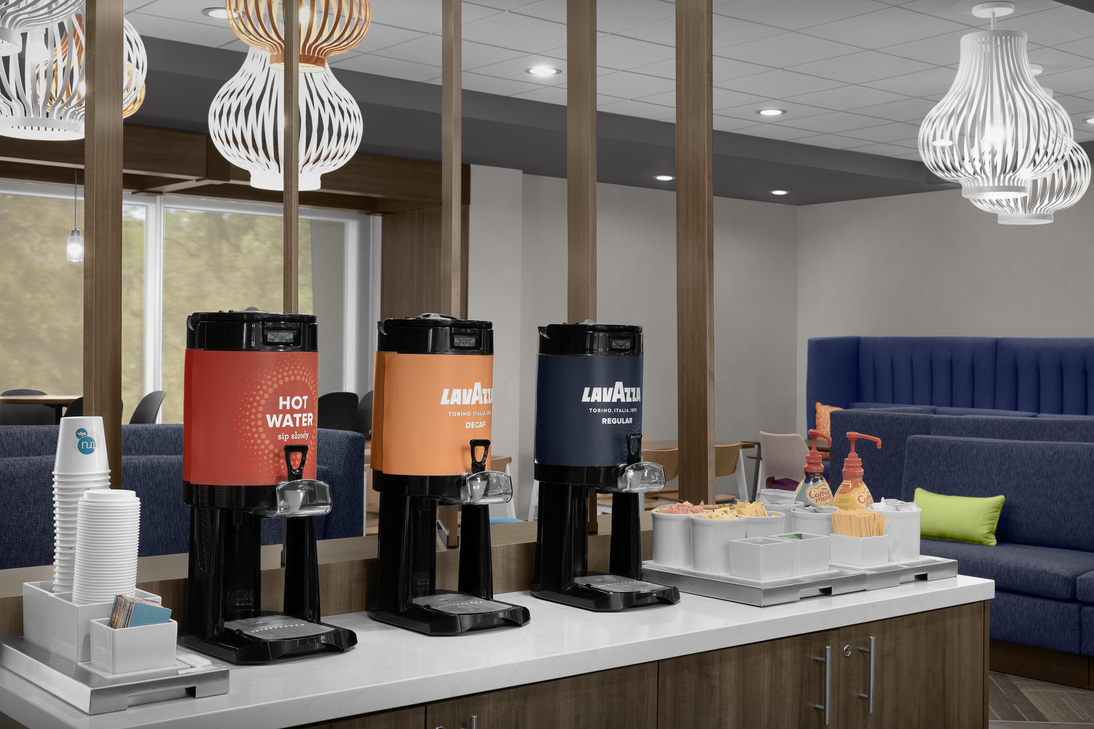 Una estación de bebidas de autoservicio con agua caliente, café regular y opciones de café descafeinado.