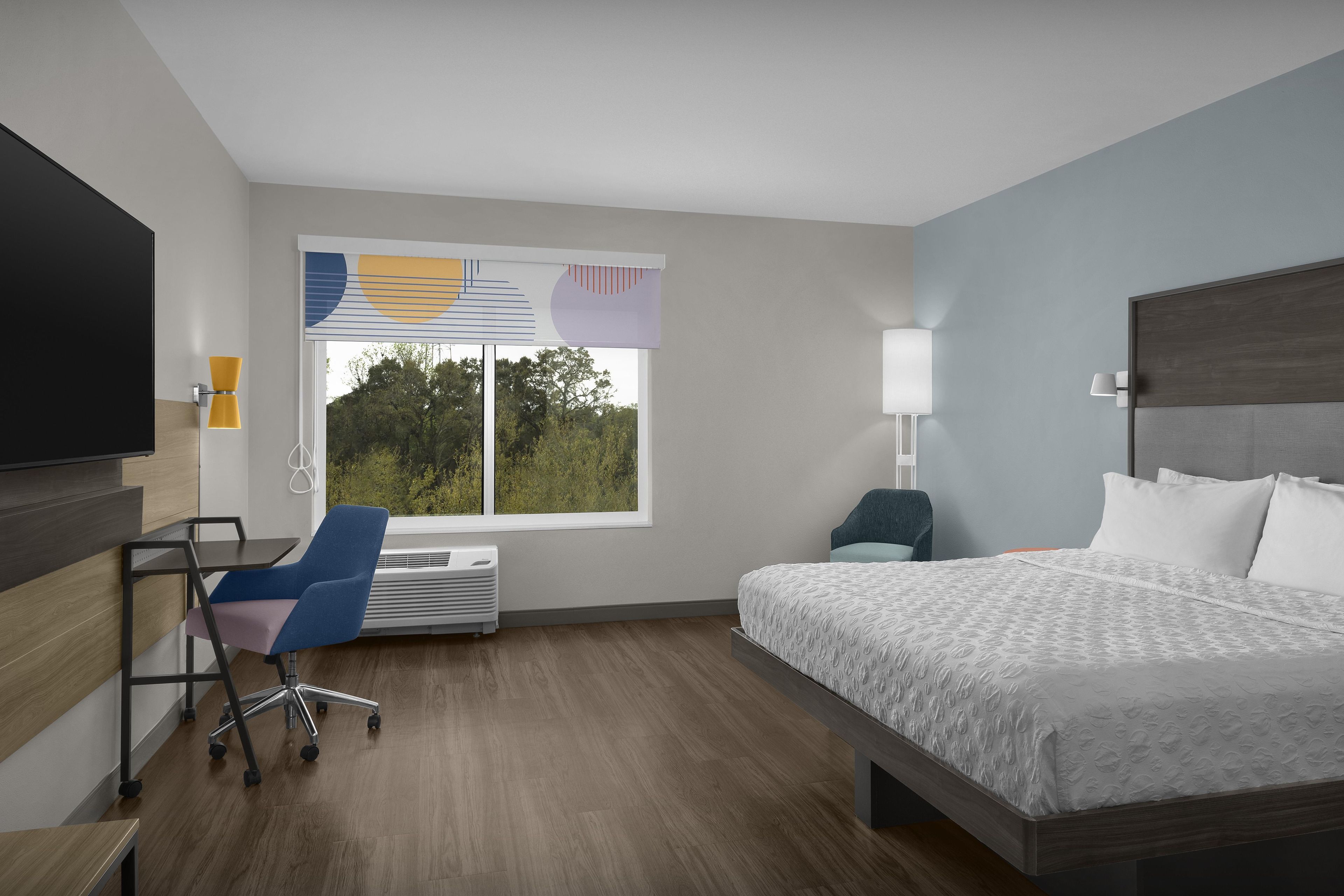 A hotel room with a bed, a desk, a chair, a TV, and a window with blinds.