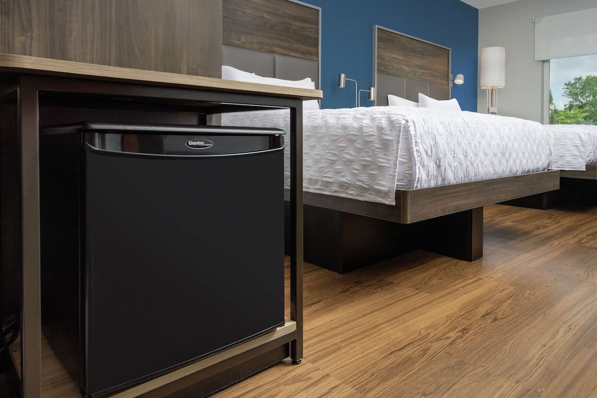 Un refrigerador compacto en una habitación de hotel con piso de madera, una cama con ropa blanca y una lámpara de pared.