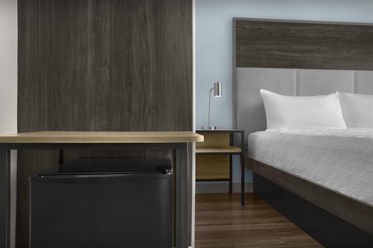 Una habitación de hotel moderna con suelo de madera, una cama con sábanas blancas, una mesita de noche, una lámpara y una pequeña mesa.