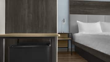 A modern hotel room with a wooden floor, a bed with white linens, a nightstand, a lamp, and a small table.