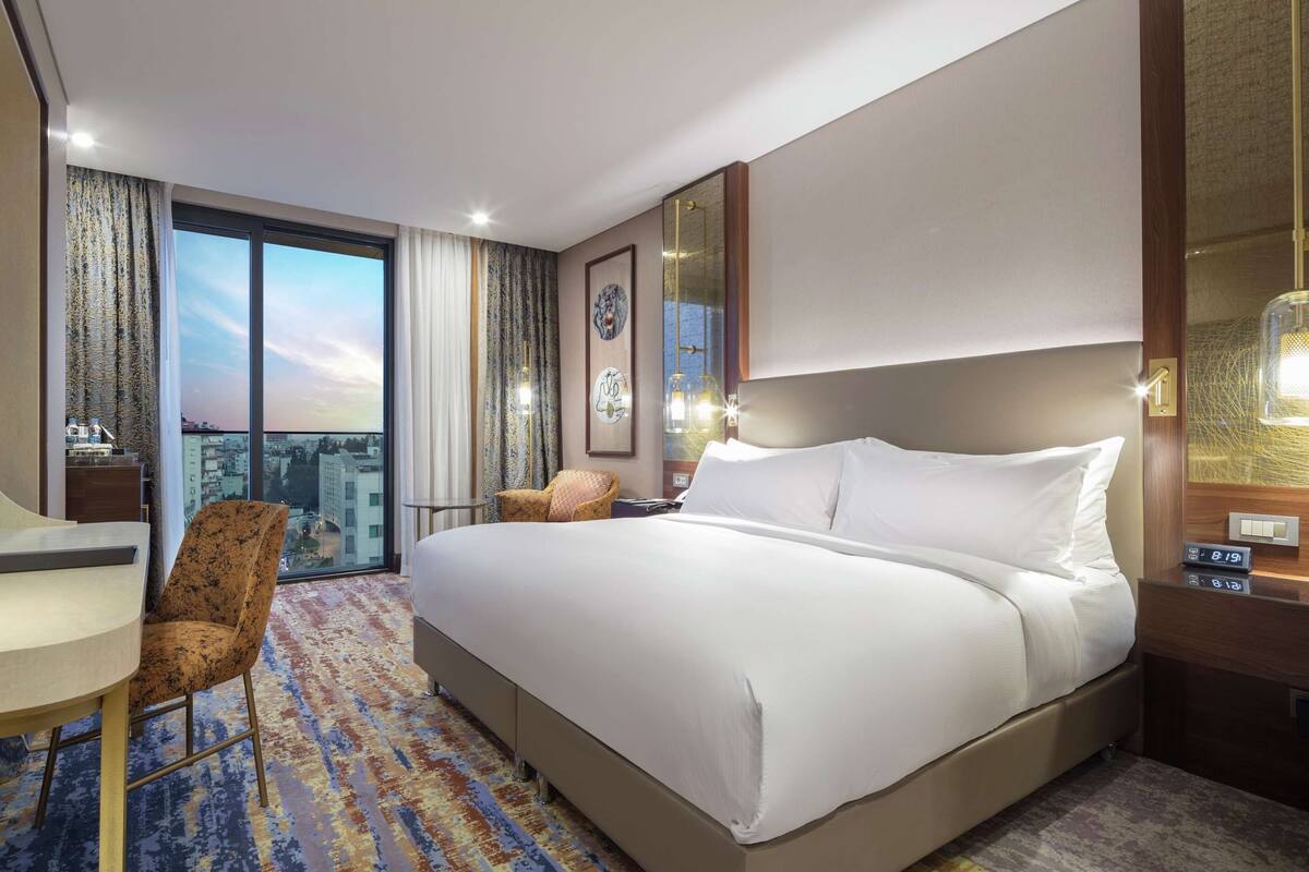 A hotel room with a large bed, a desk, a chair, a TV, and a balcony with a city view.