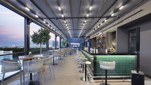 Un bar moderno con mostrador de azulejos verdes, varios taburetes y vistas panorámicas de la ciudad a través de grandes ventanales.