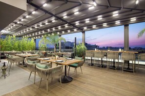 Un área de comedor al aire libre con varias mesas y sillas, ventanas amplias que ofrecen vista a la ciudad e iluminación empotrada en el techo.