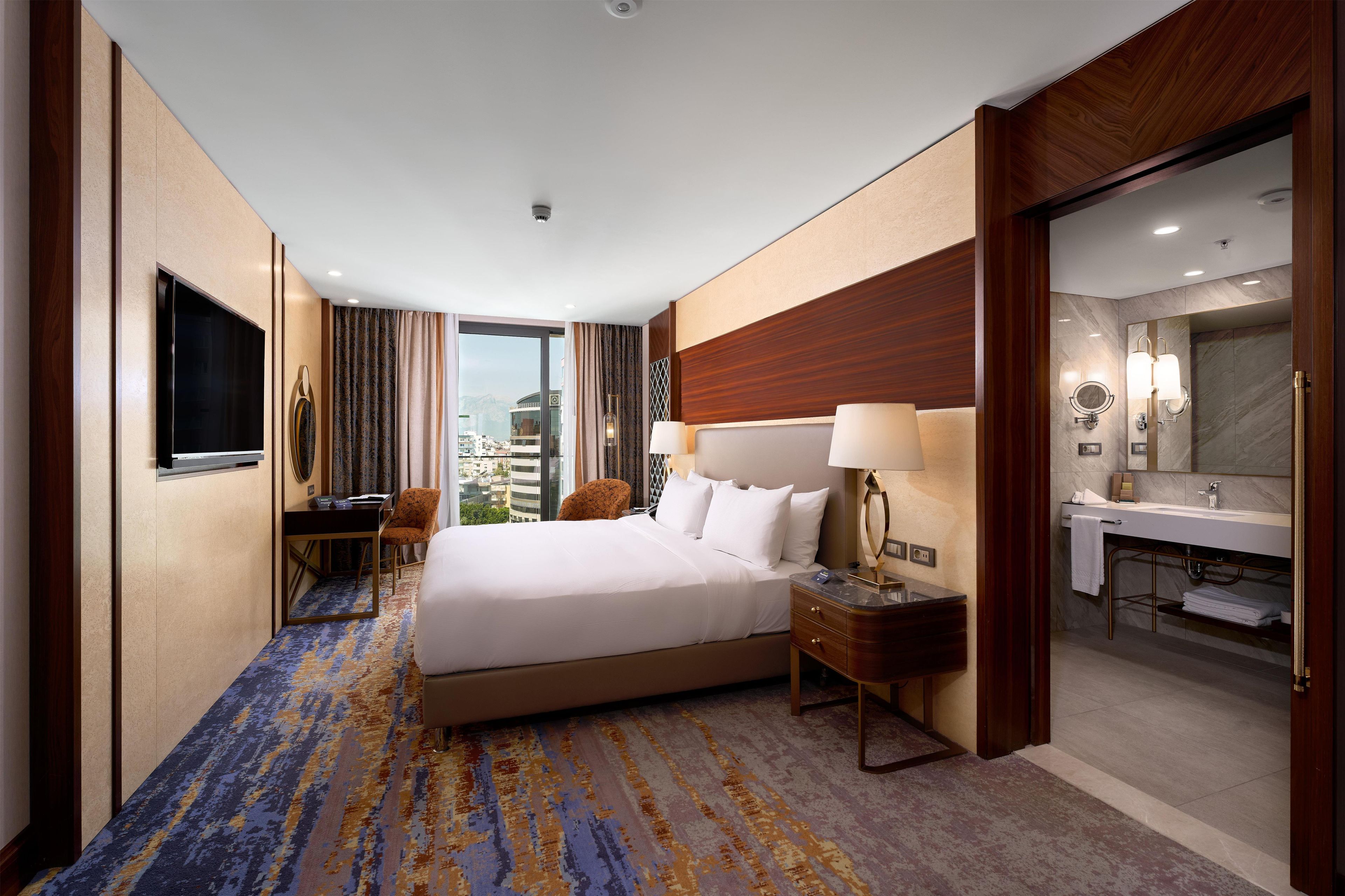 A hotel room with a large bed, a desk, a chair, a television, and a bathroom with a mirror and sink.