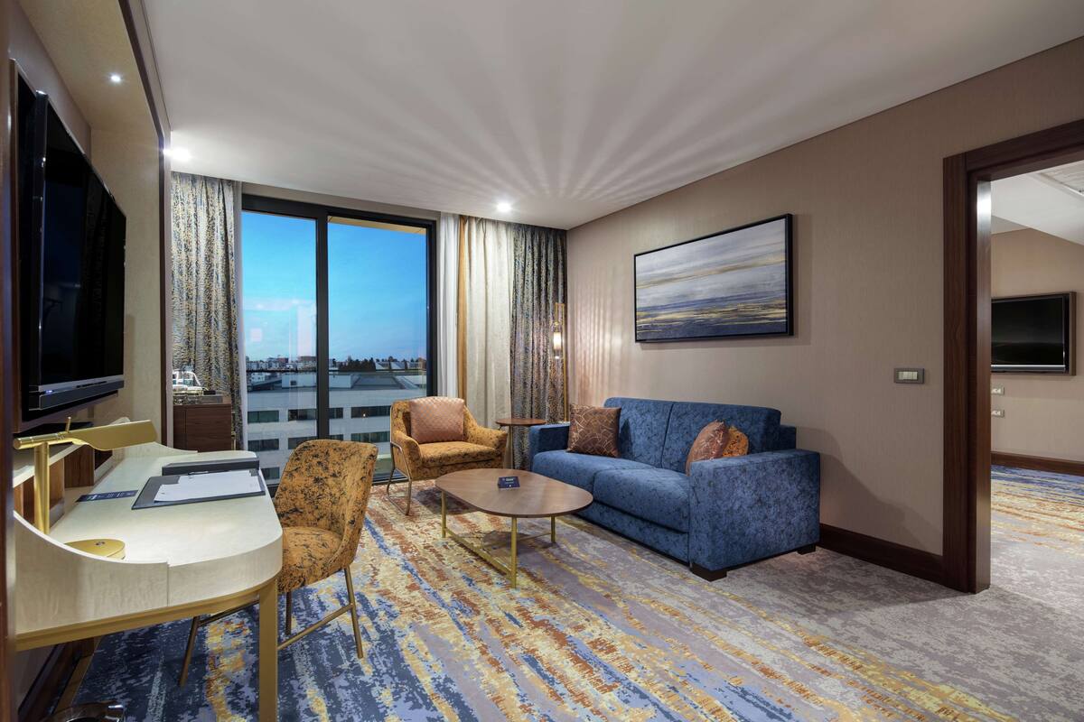 A hotel room with a large window, a sofa, a coffee table, a desk with a chair, and a television.