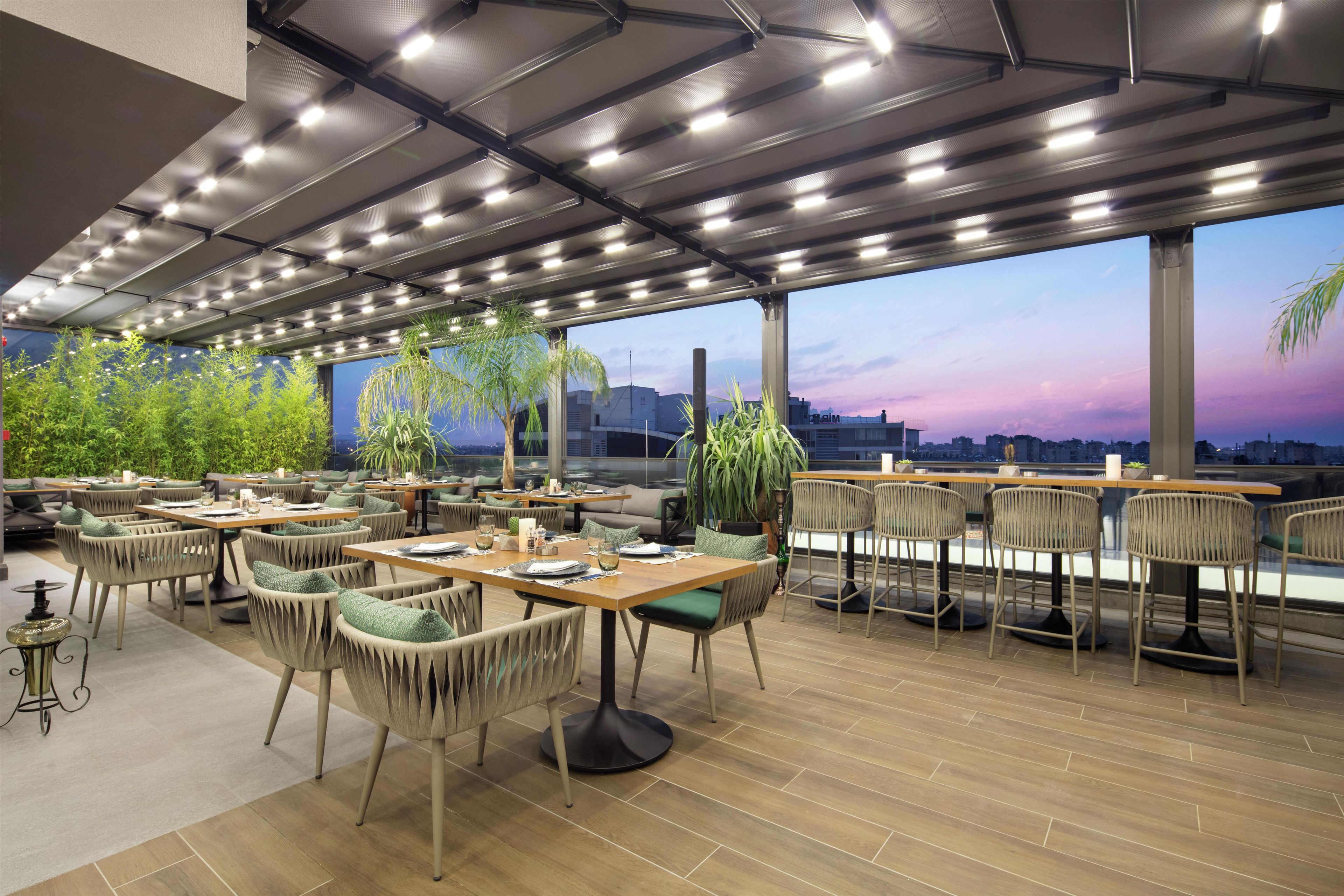 Un área de comedor al aire libre con varias mesas y sillas, grandes ventanales y vistas a un horizonte urbano.
