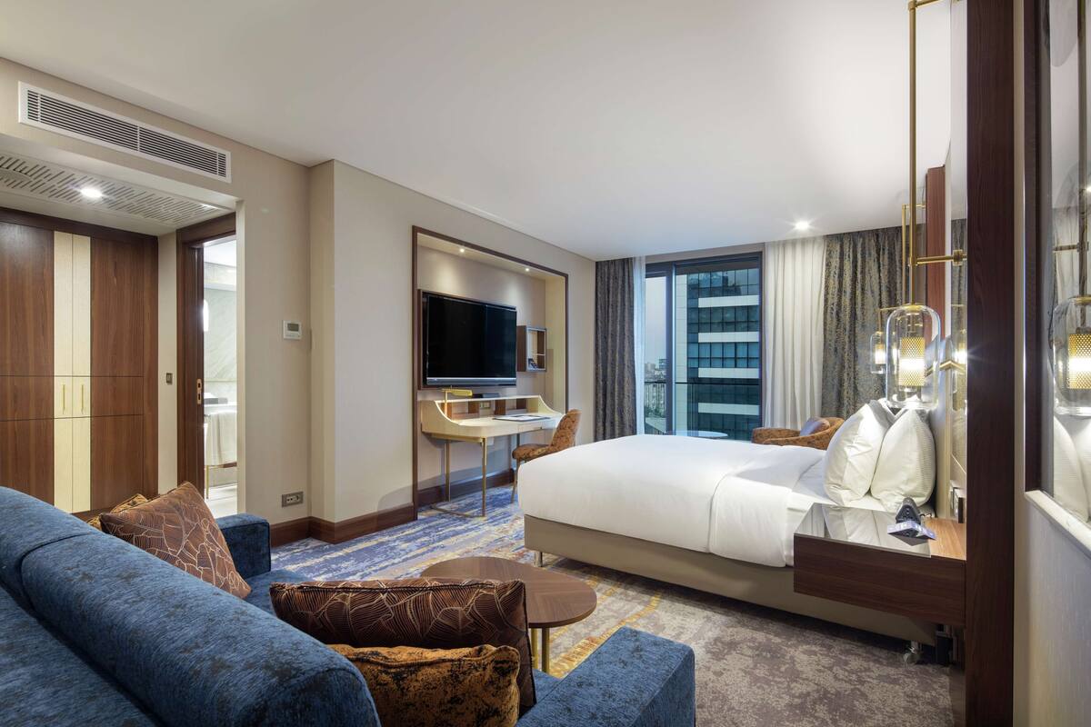A hotel room with a large bed, a sofa, a desk with a TV, and a view of the city through the window.