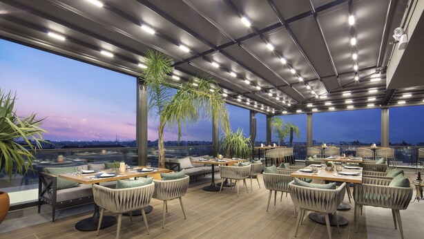 Un espacio exterior para comer con mesas y sillas, ventanas amplias que ofrecen vista de la ciudad y un techo con iluminación empotrada.