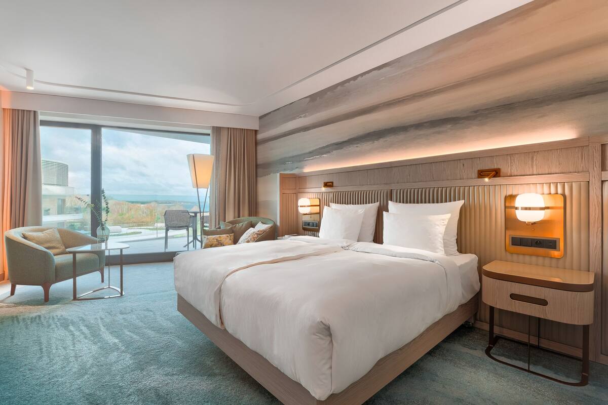 Una habitación de hotel moderna con una cama grande, una zona de estar con una silla y una mesa, y vistas al océano a través de amplios ventanales.