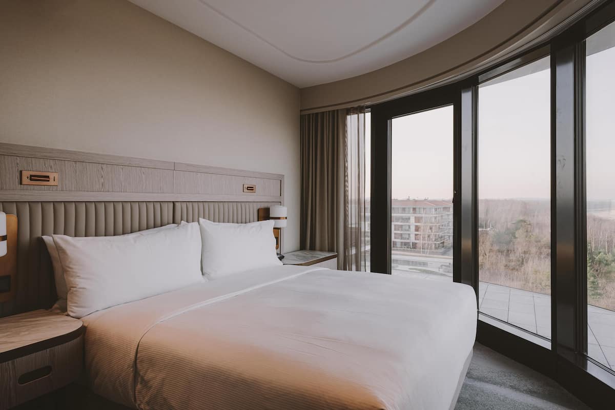 Habitación de hotel con una cama grande, una mesita de noche, una lámpara y vistas a la ciudad a través de amplios ventanales.