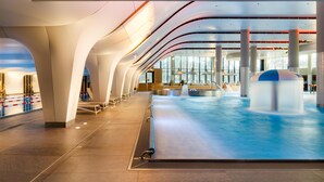 Una piscina interior moderna con techo curvo, amplias ventanas y un elemento acuático central.