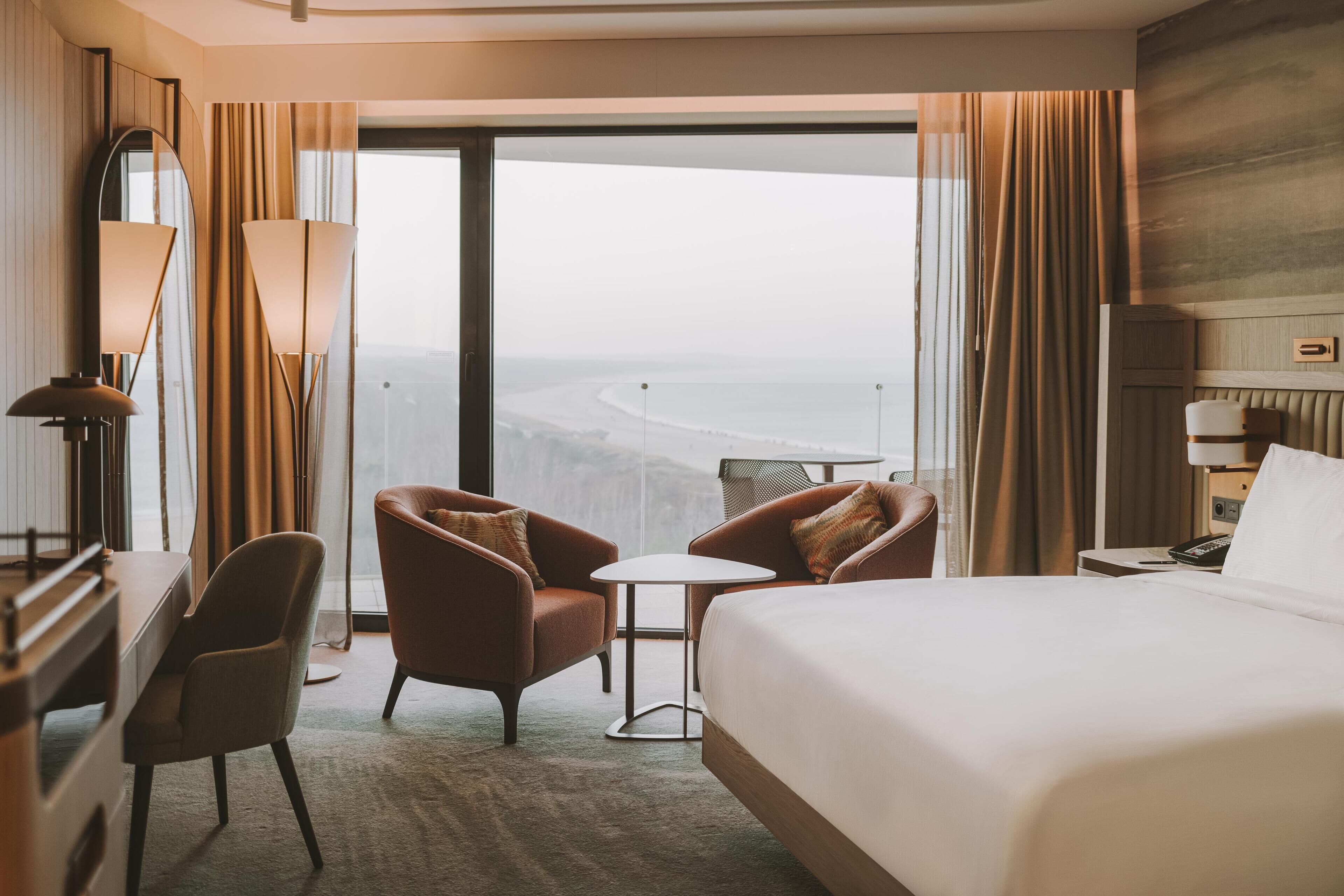 Een hotelkamer met een groot bed, twee fauteuils, een kleine tafel, een bureau, een lamp, een spiegel en uitzicht op de oceaan.