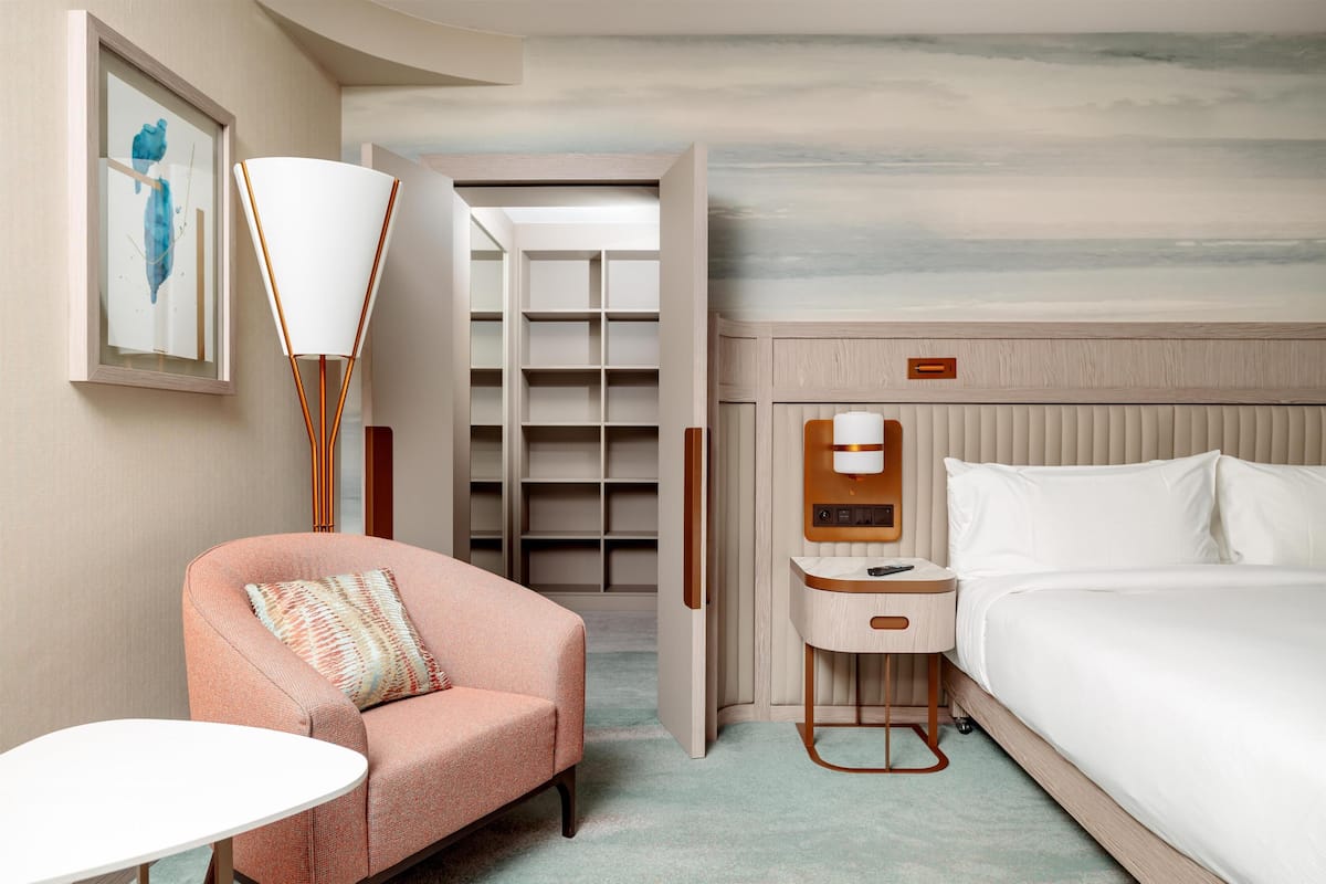 Habitación de hotel con cama, una silla, una mesita, lámpara, armario y un cuadro en la pared.