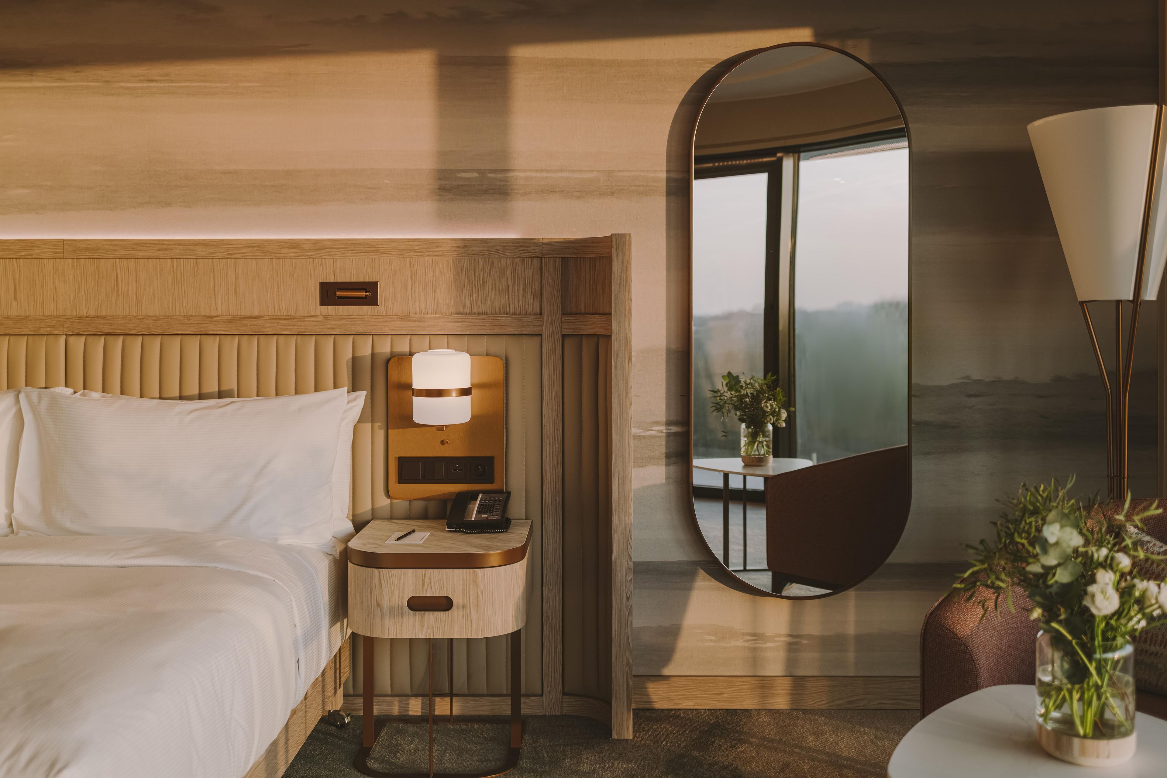 A hotel room with a bed, a bedside table with a lamp, a mirror, and a view of the outdoors.