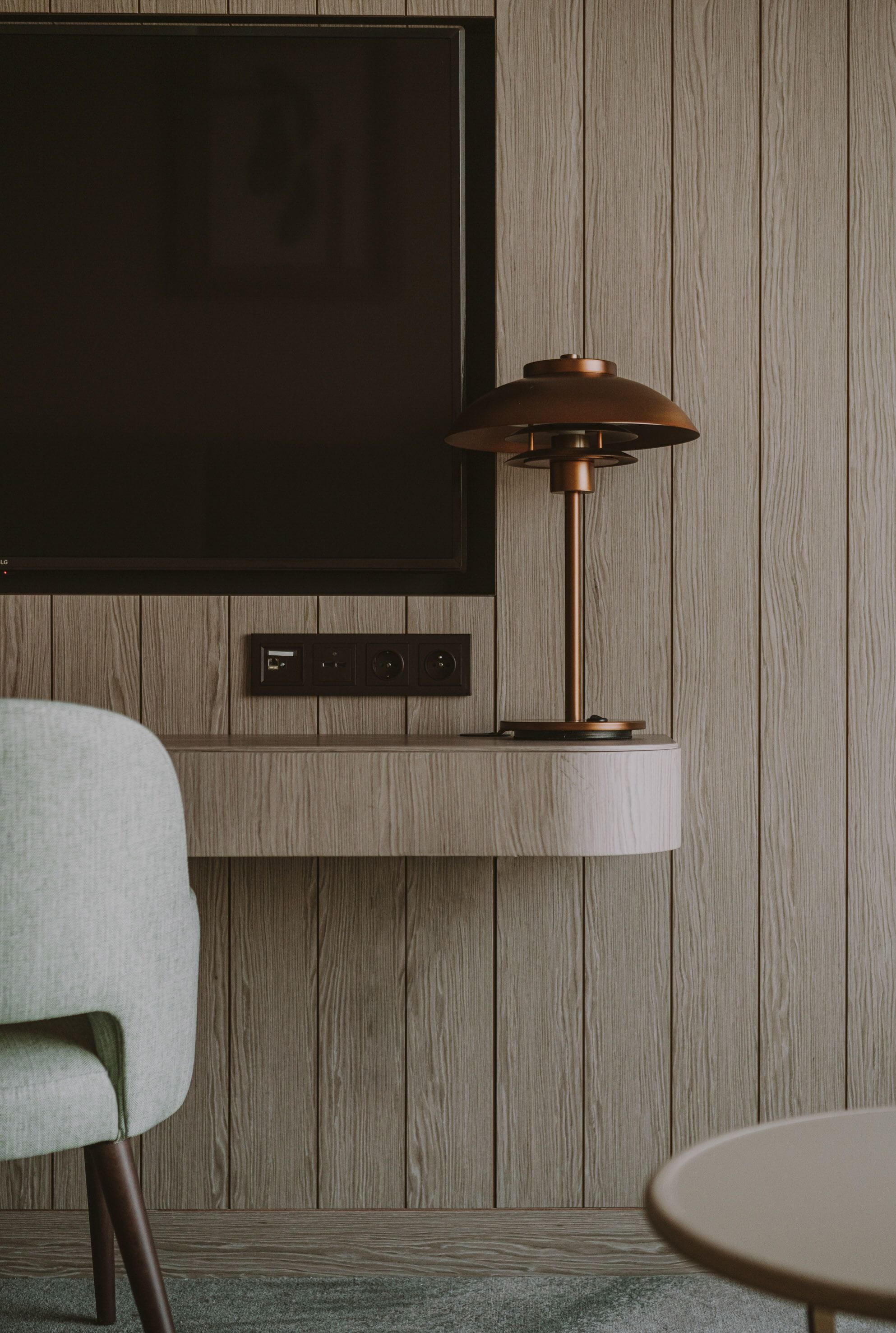 A modern room with a mounted flat-screen TV, a wall-mounted lamp, and a seating area with a chair and a small round table.