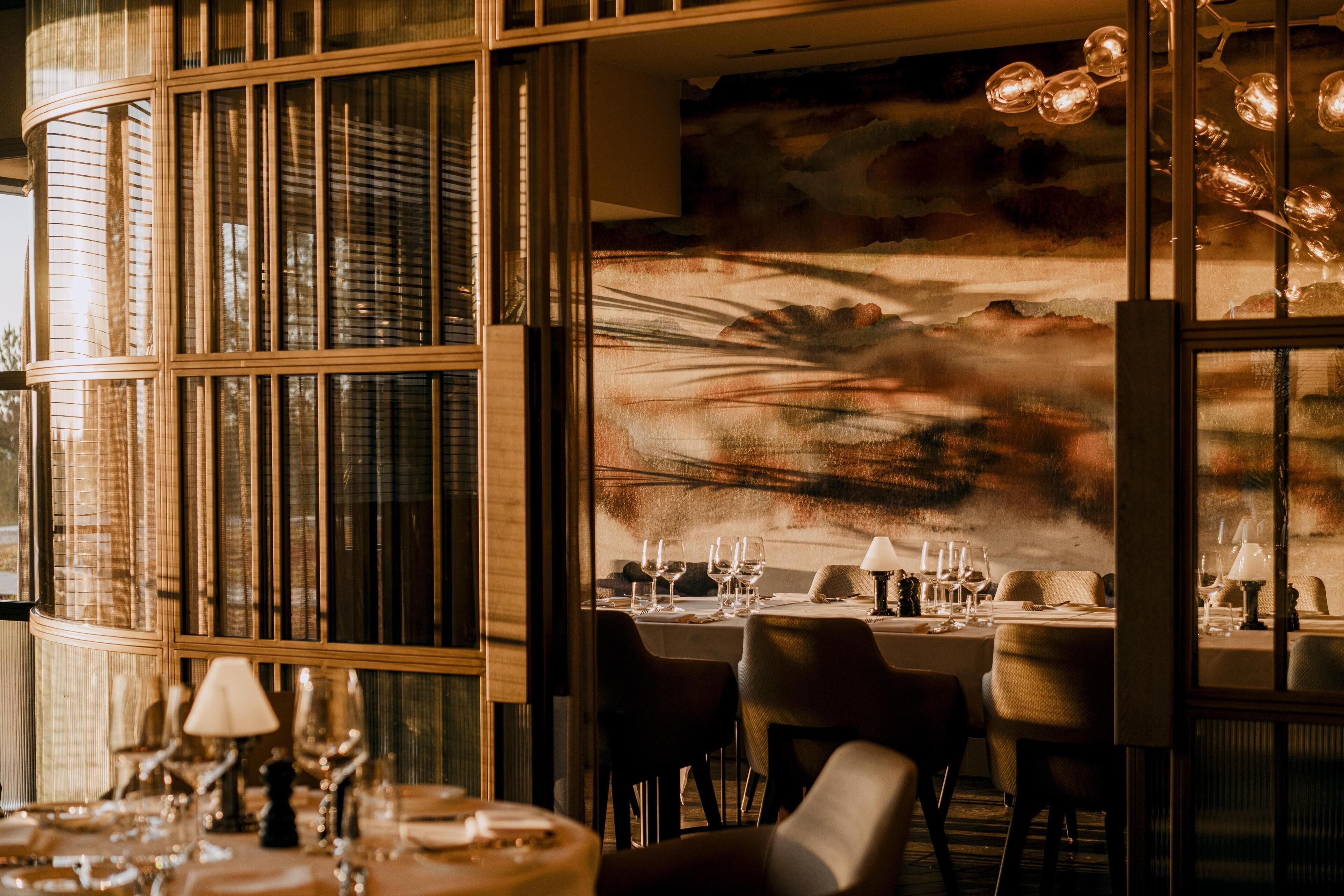 A restaurant interior with large windows, tables set for dining, and a mural on the wall.