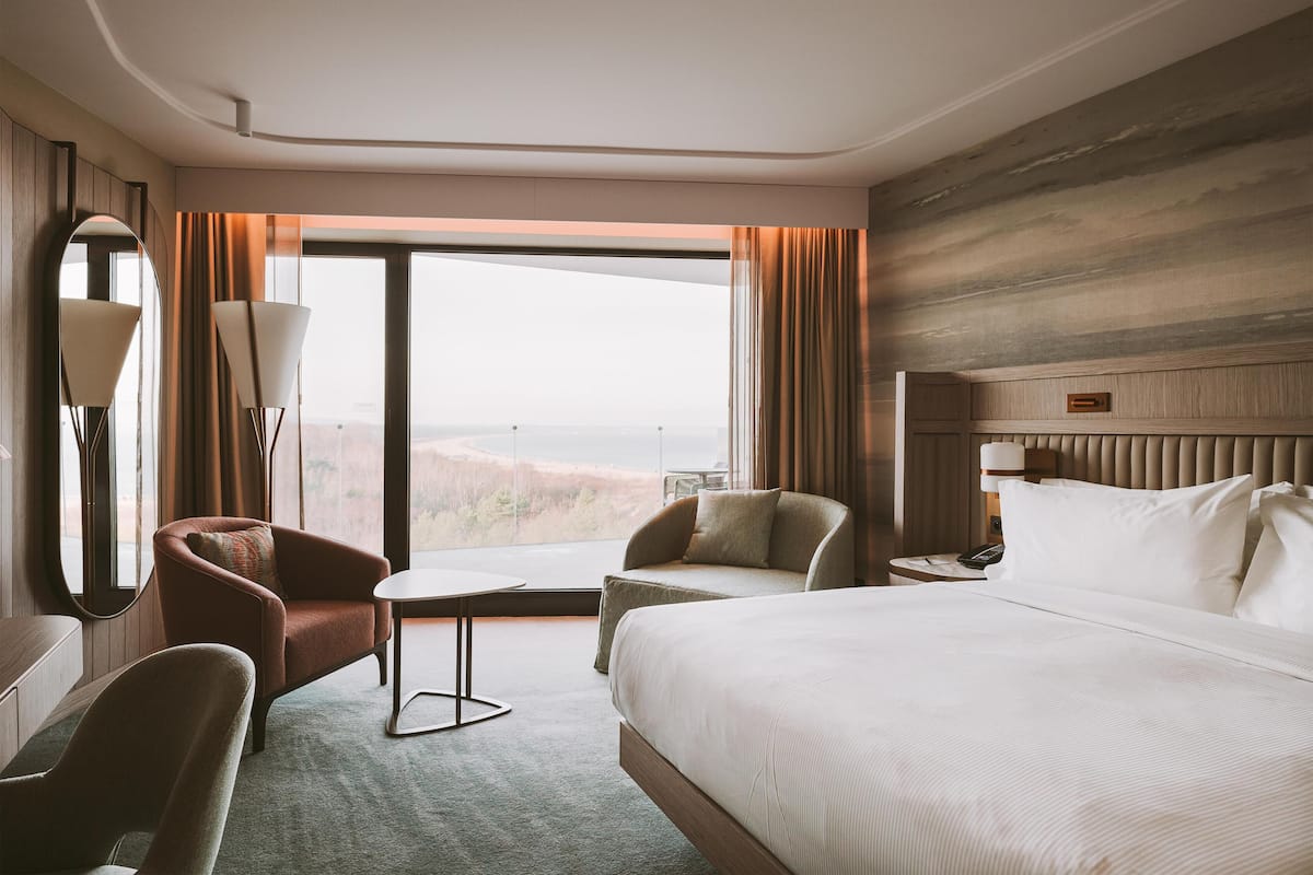 Habitación de hotel con una cama grande, dos sillones, una mesita, un escritorio, un espejo y vistas al exterior.