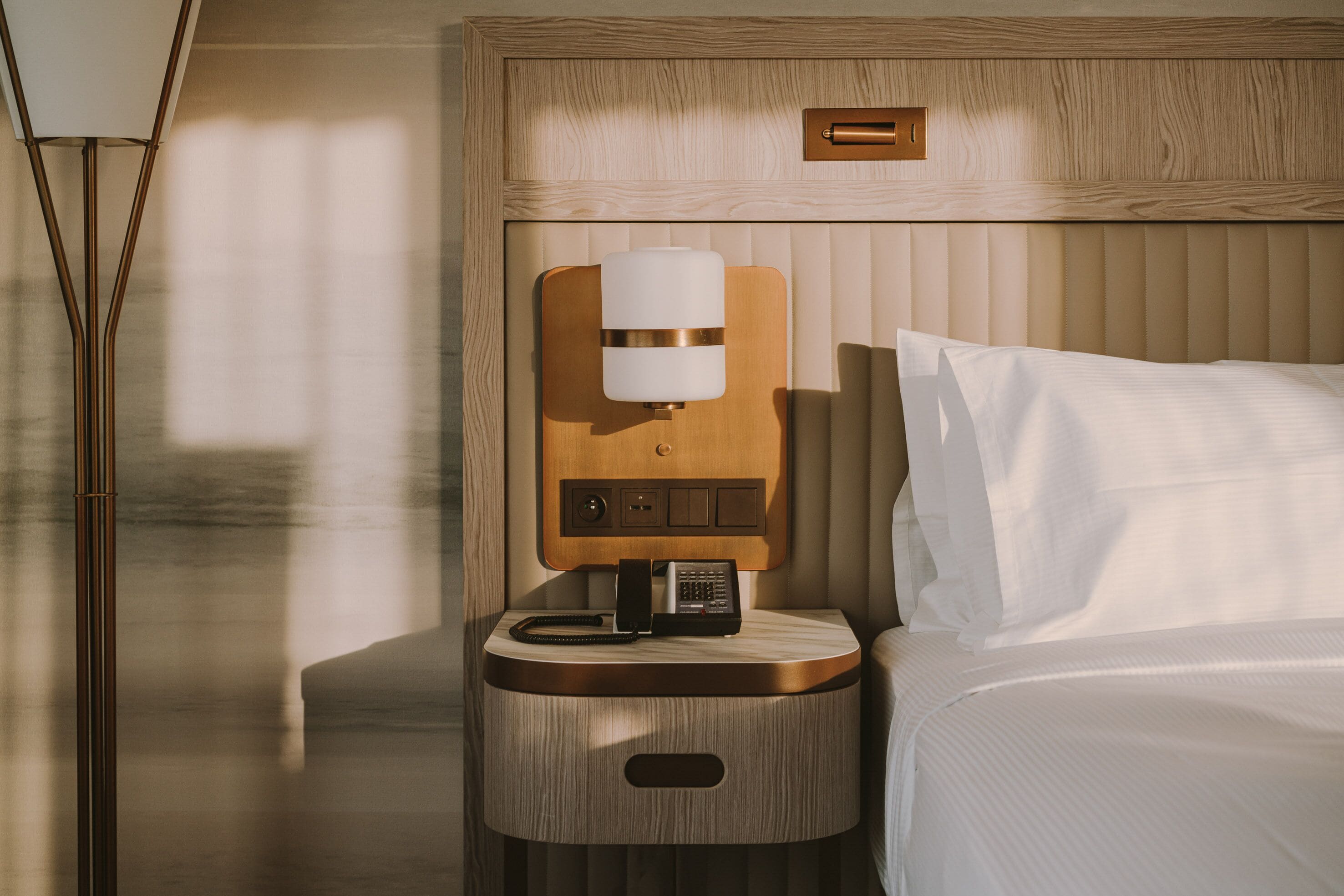 Une chambre d’hôtel bien rangée, avec une table de chevet en bois, une lampe de chevet, un téléphone et un lit recouvert de draps blancs.