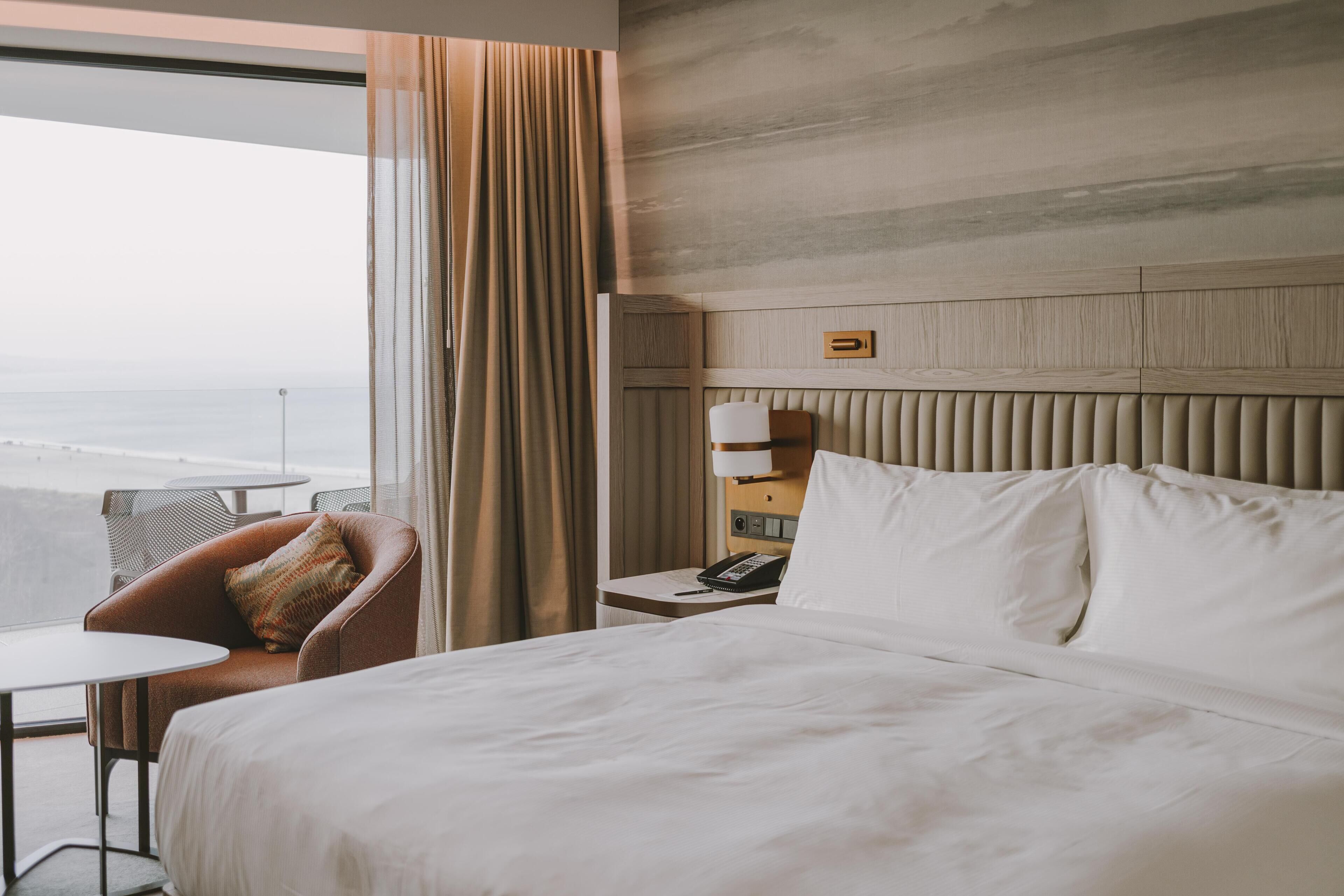 A hotel room with a bed, a chair, a small table, a nightstand, a lamp, and a view of the ocean.