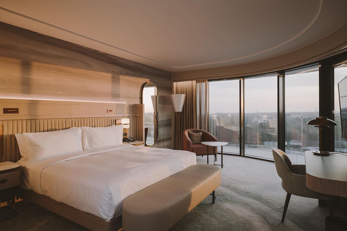 Una habitación de hotel moderna con una cama grande, una mesita pequeña, una silla, un espejo y un amplio ventanal con vistas a la ciudad.