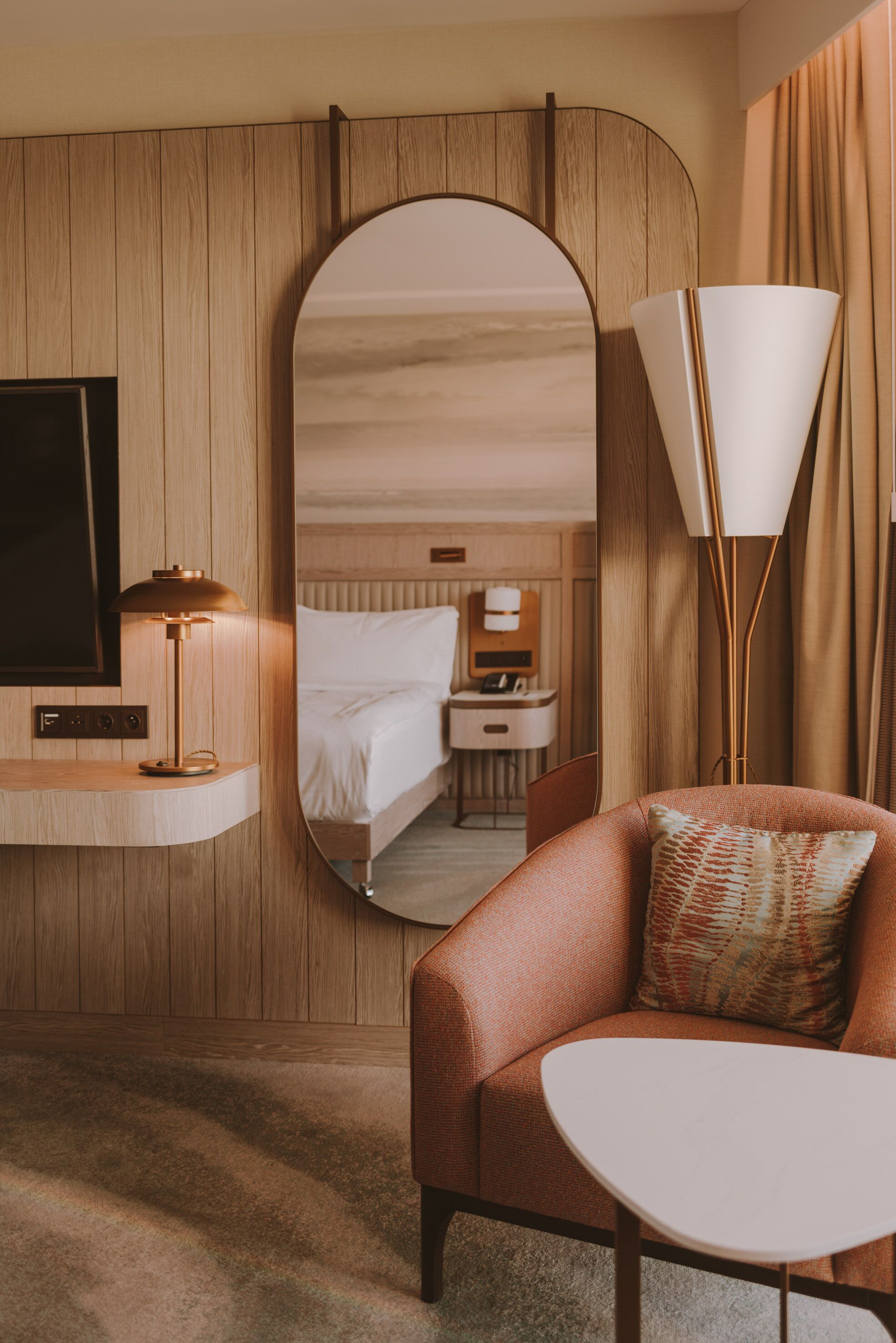 A hotel room with a wooden panel wall, a large mirror, a bed, a desk with a lamp, and a seating area with a chair and table.