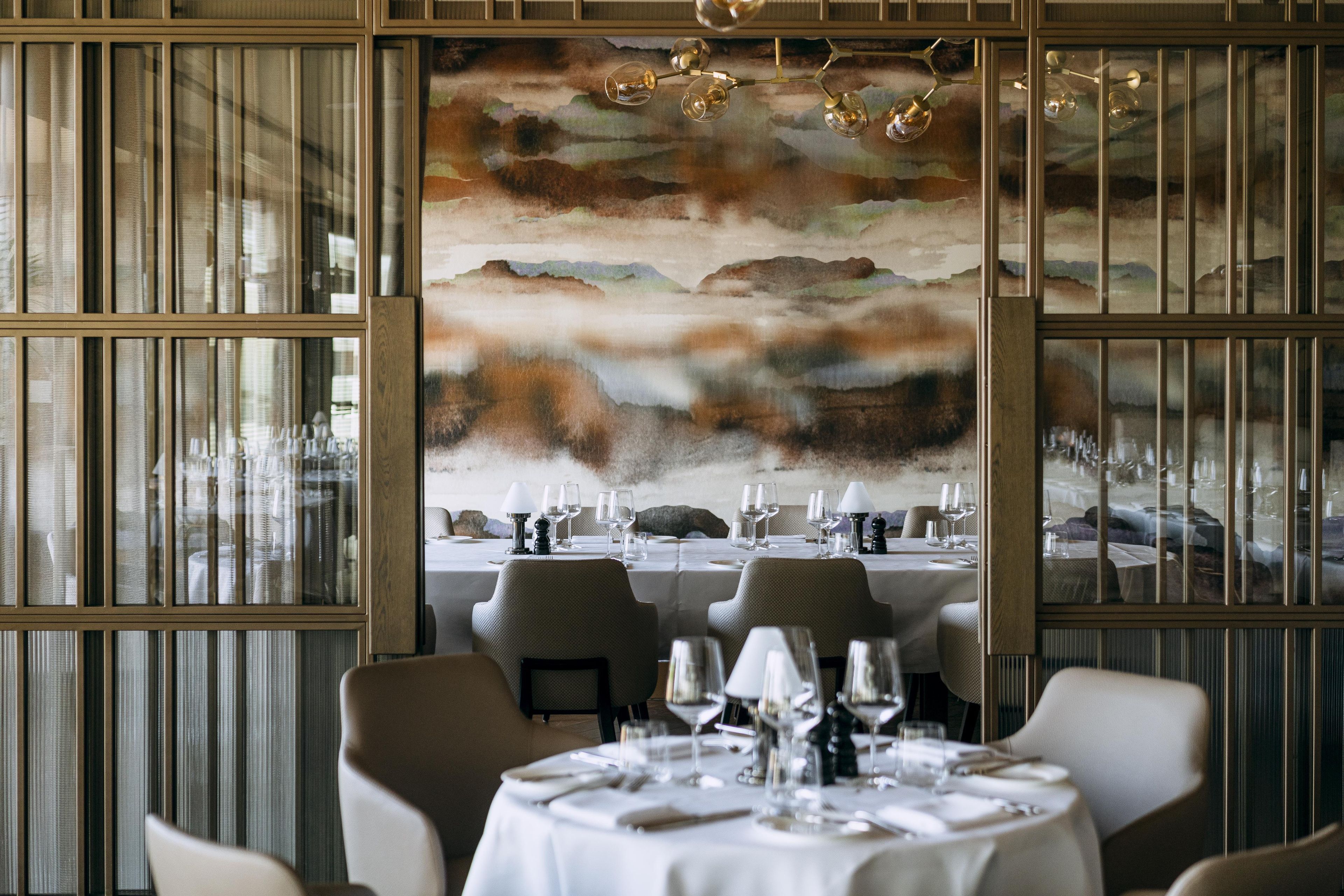 A dining area with tables set for a meal, a large abstract painting on the wall, and a view through the window.