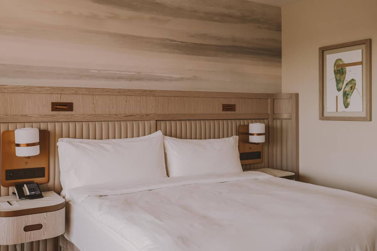 Una habitación de hotel con una cama grande, dos mesitas de noche con lámparas, un cabecero de madera y una obra de arte colgada en la pared.