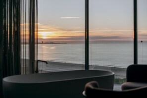 A room with a large window overlooking a beach at sunset.