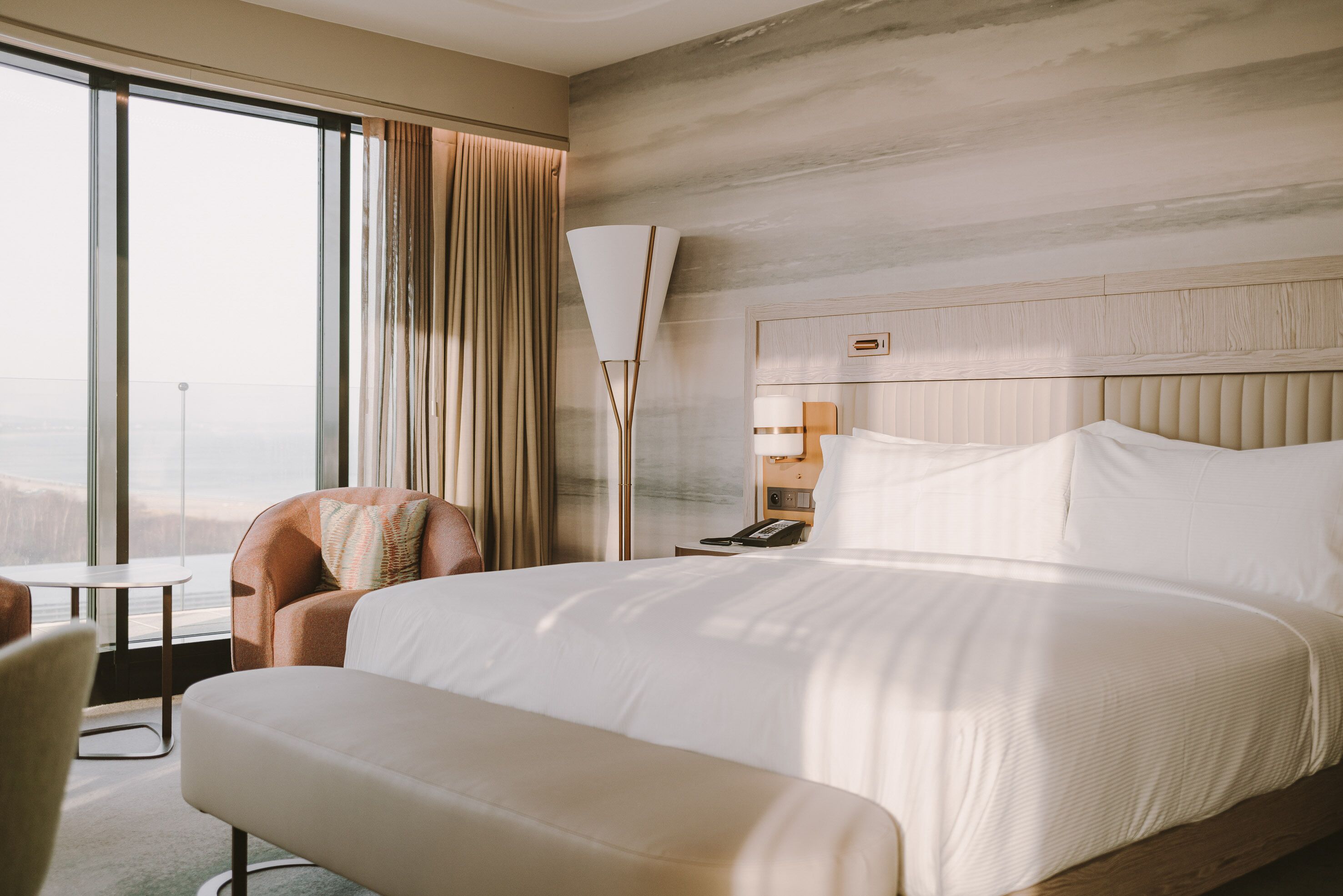 A hotel room with a large bed, a sitting area with a chair, a small table, and a view of the city through large windows.