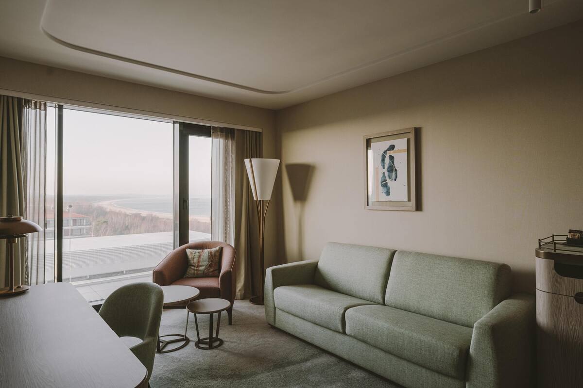 Habitación de hotel con un sofá verde, una mesita redonda pequeña, una silla, una lámpara y un ventanal con cortinas.