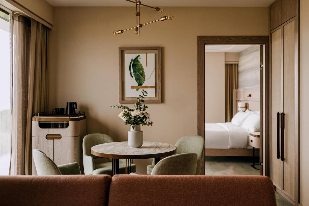 Una habitación de hotel con una cama, una mesa de comedor con sillas, una cafetera, un florero con flores y una obra de arte enmarcada en la pared.