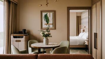 Quarto de hotel com cama, uma mesa de jantar com cadeiras, máquina de café, um vaso com flores e uma obra de arte emoldurada na parede.