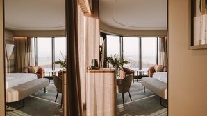 Habitación de hotel moderna con techo curvo, ventanas amplias, una mesa de madera en el centro, dos sofás y un escritorio pequeño.