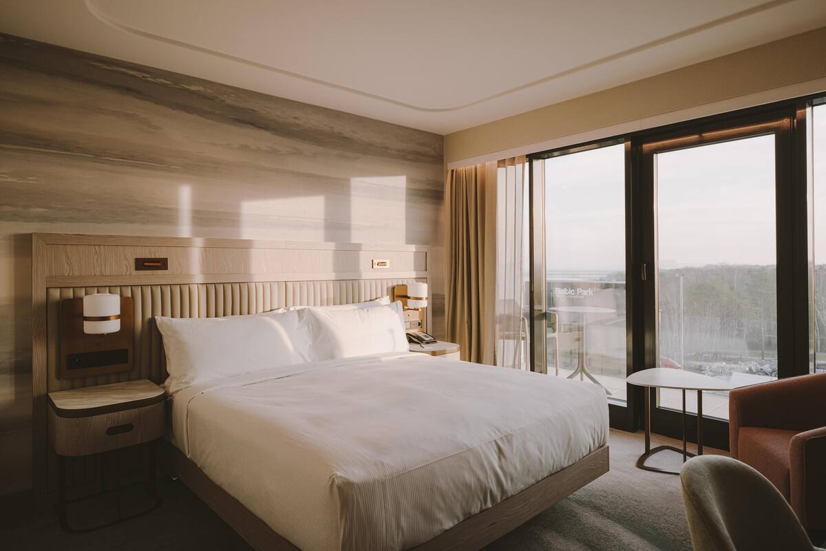 Una habitación de hotel con una cama grande, una mesita de noche, un escritorio, una silla y vistas a un paisaje urbano a través de un ventanal amplio.