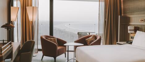 Habitación de hotel con cama, dos sillones, una mesita, un escritorio con lámpara y vista al océano.