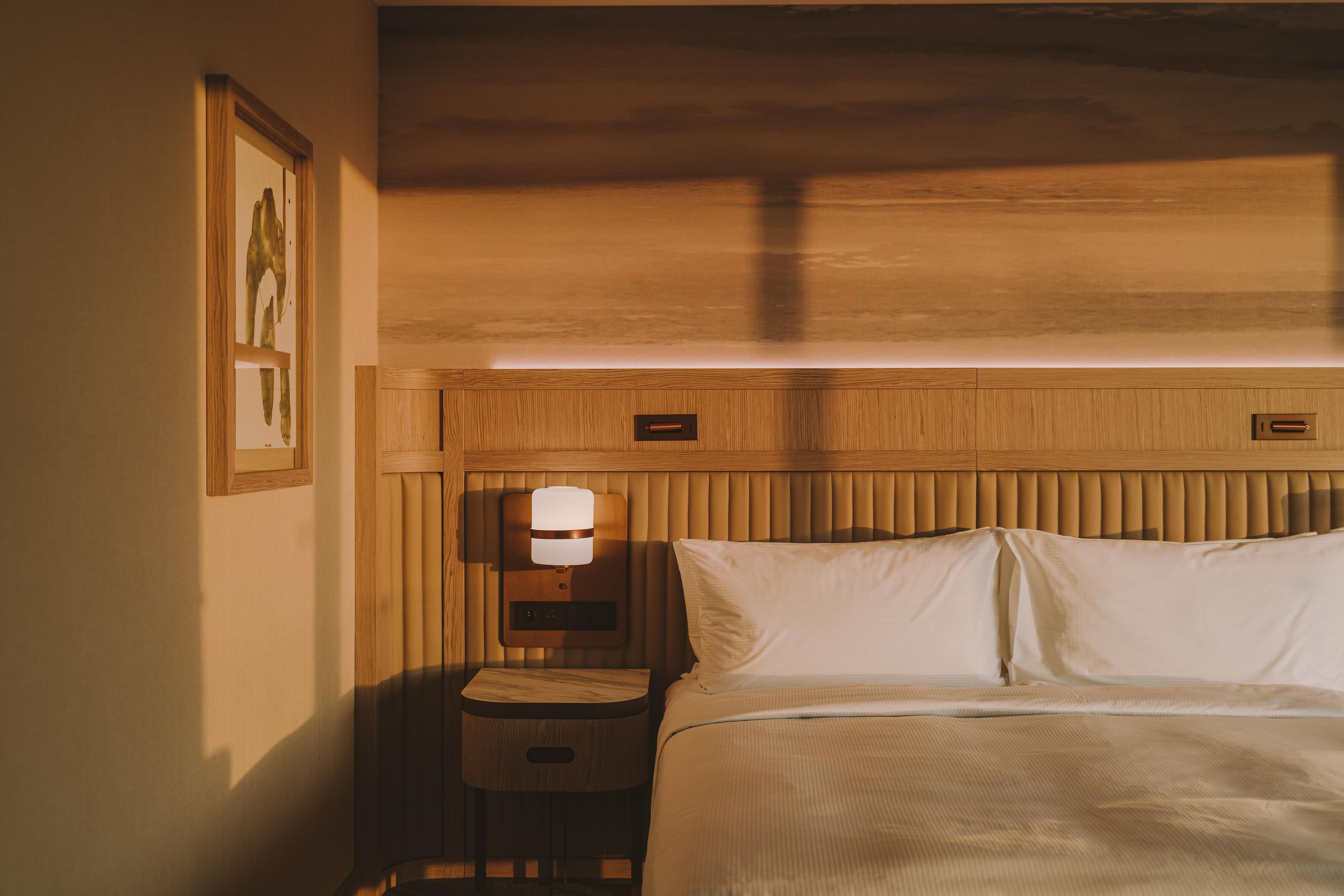 A hotel room with a bed, a nightstand, a wall-mounted lamp, and a framed picture.