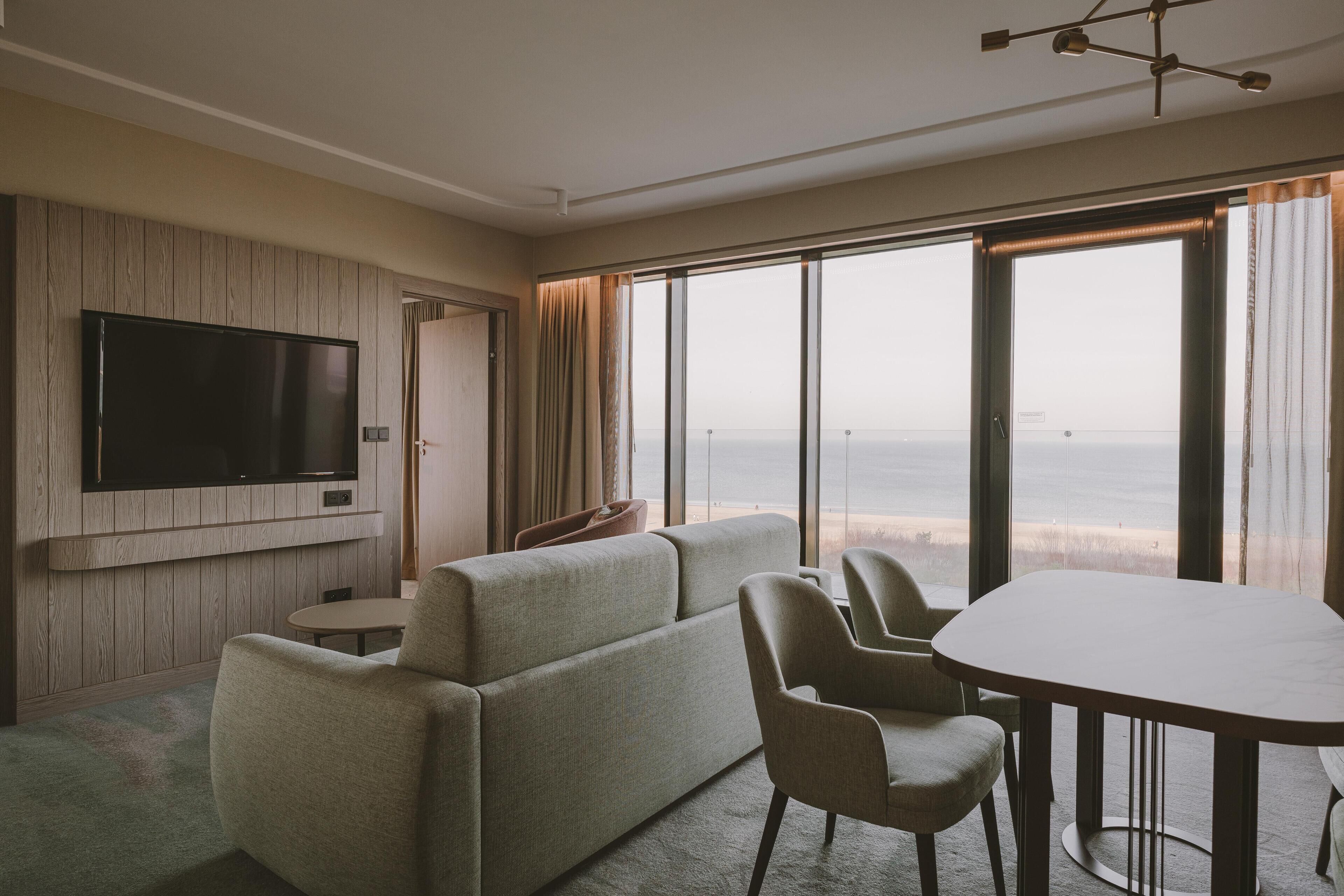 A hotel room with a large TV, a sofa, a dining table, and a view of the ocean.