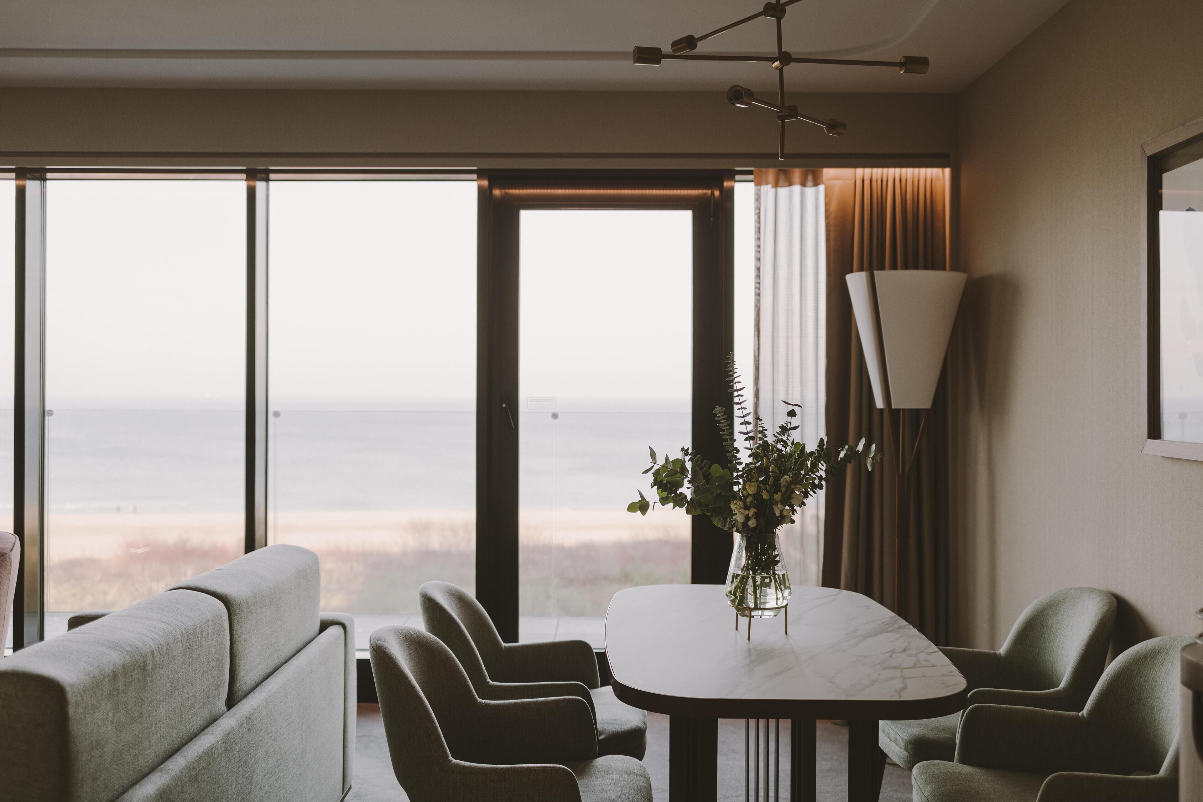 A modern dining area with a large table, chairs, a vase with flowers, and a view of the ocean.