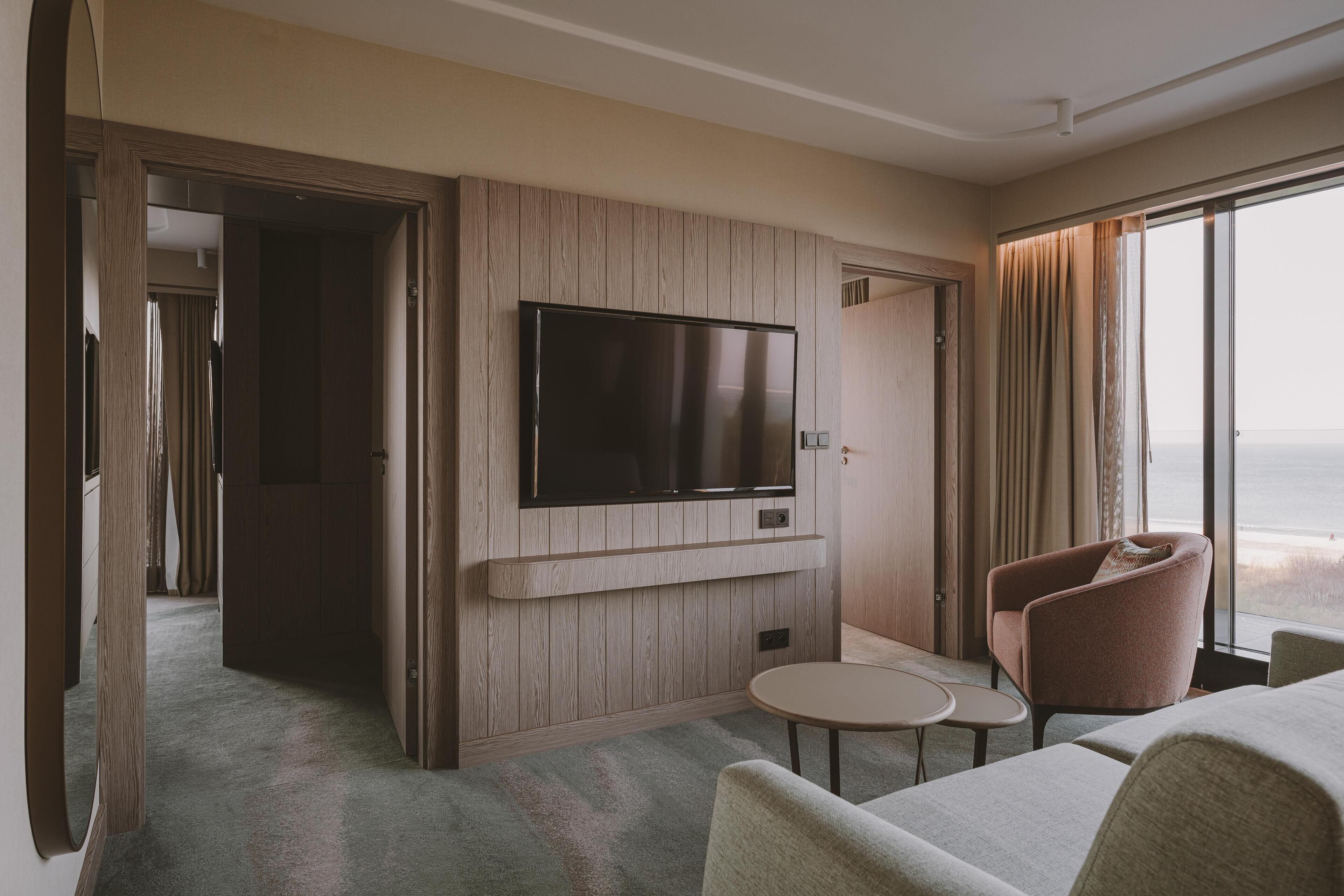 Ein Hotelzimmer mit an der Wand montiertem Fernseher, einem kleinen Tisch, einem Stuhl und Meerblick.