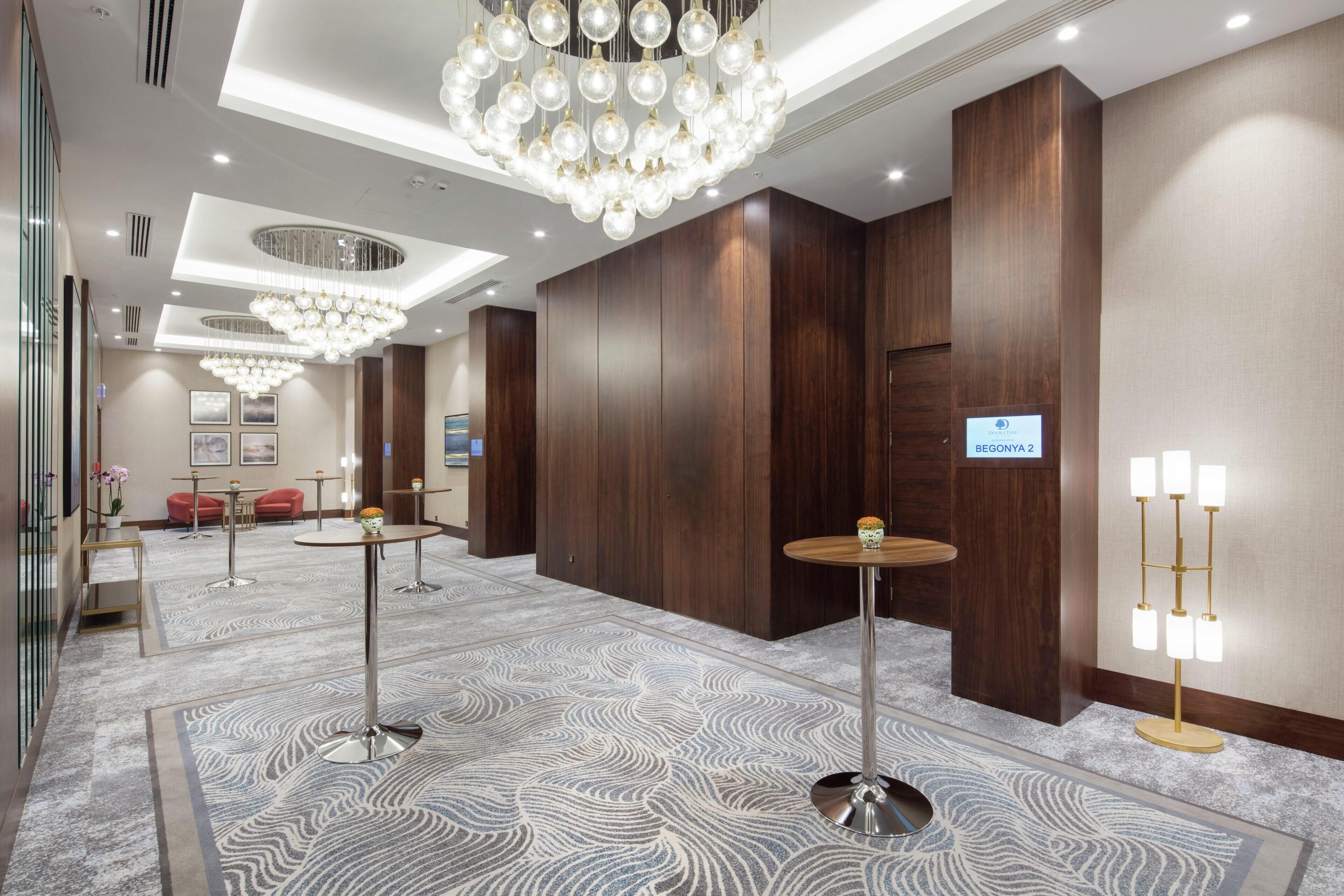 A hotel lobby with a chandelier, round tables, a reception desk, and a patterned carpet.