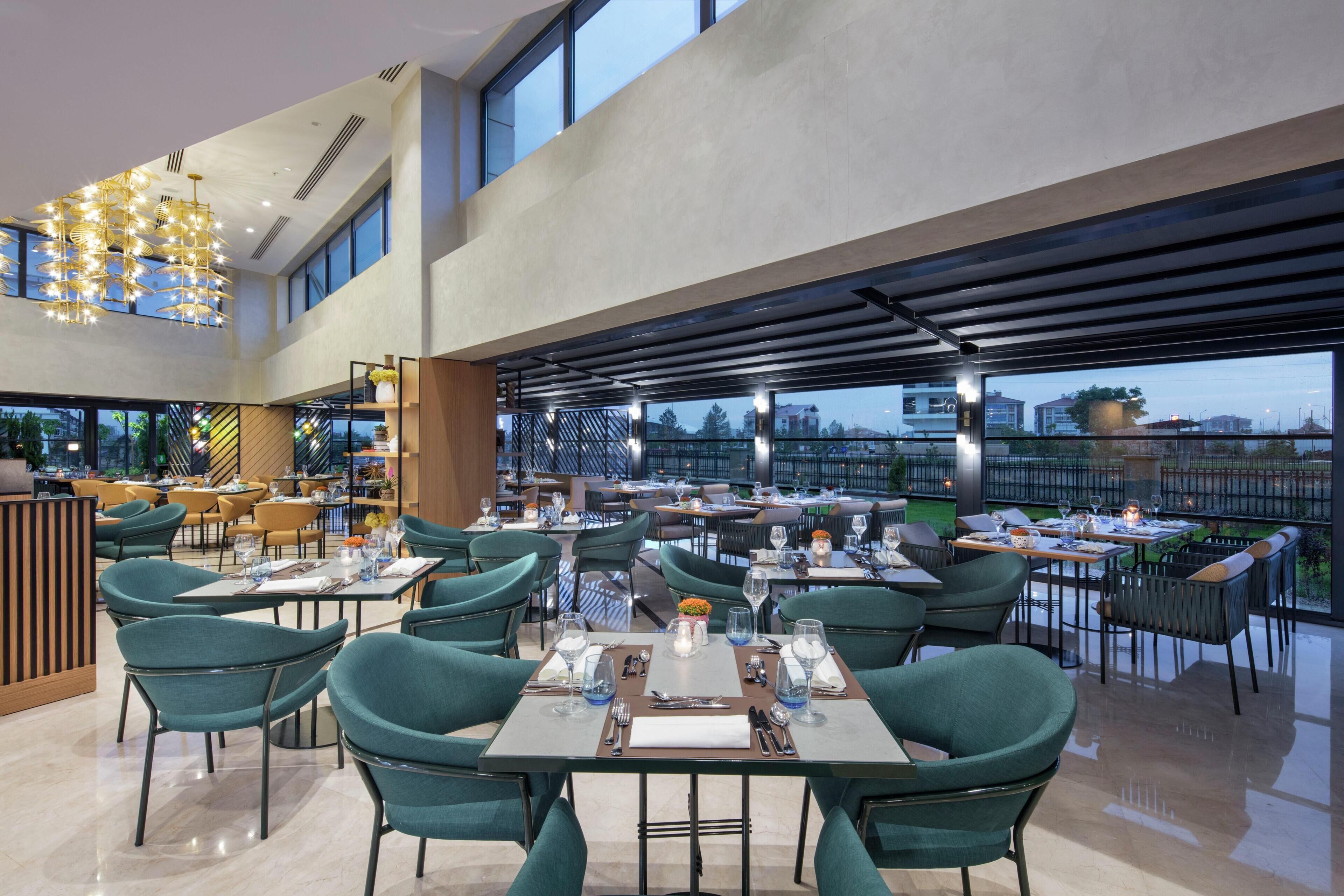 A modern restaurant with multiple dining tables and chairs, large windows, and a high ceiling with a unique light fixture.