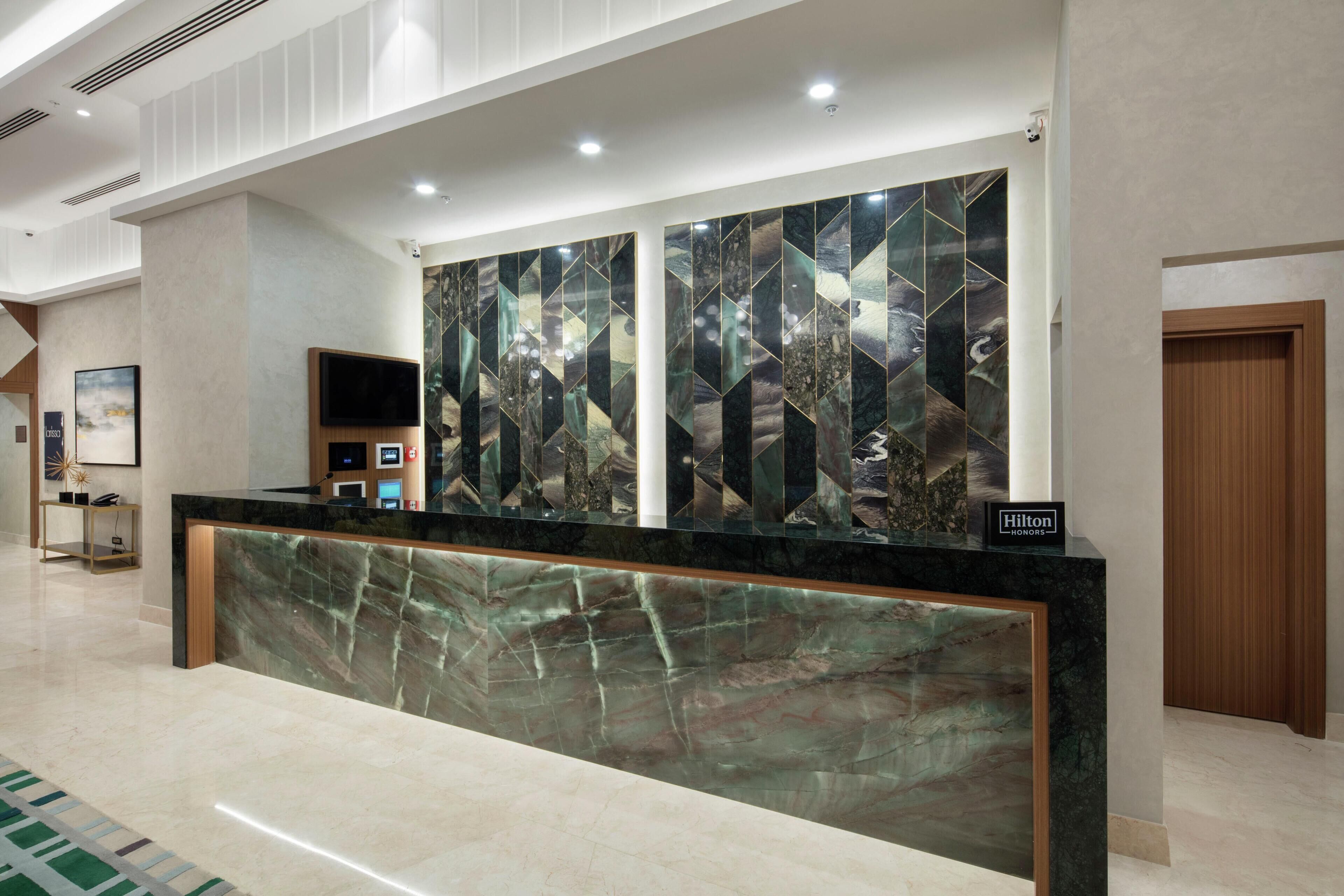 A hotel reception desk with a marble counter, a large wall art piece, a television, and a sign indicating Hilton Honors.