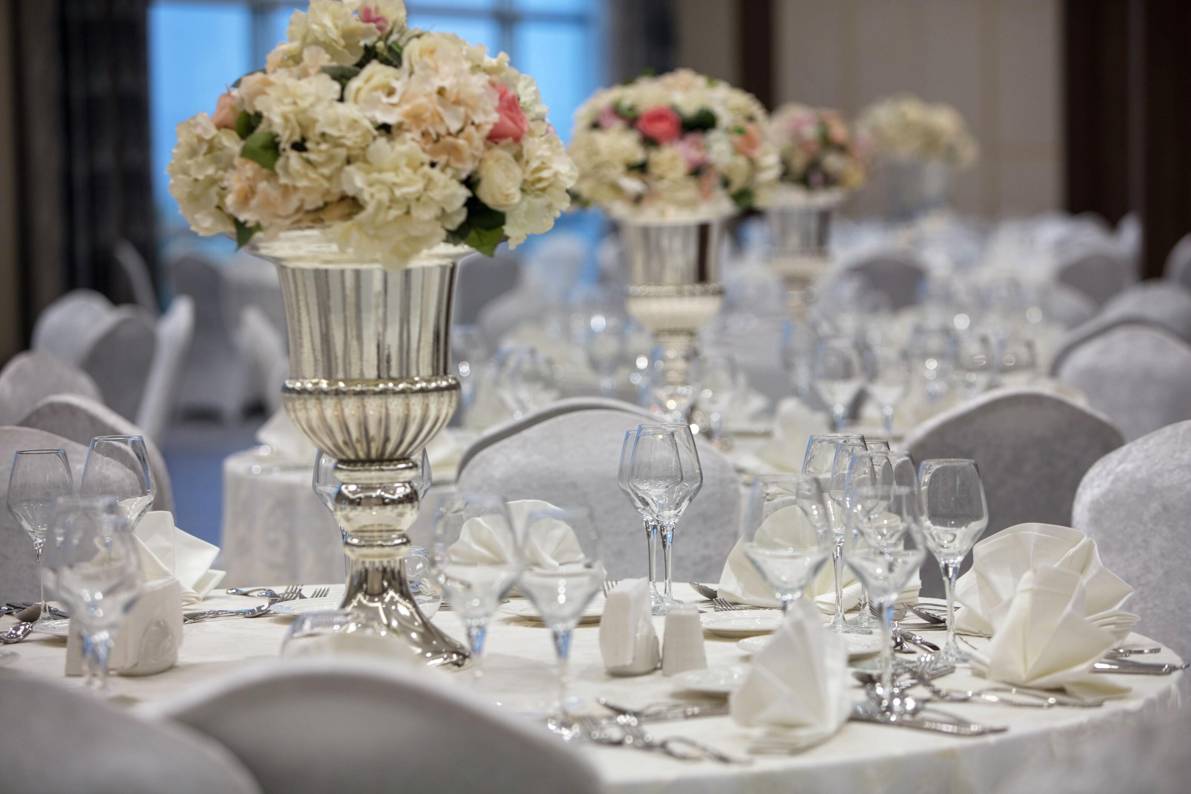 Una mesa de banquete dispuesta con manteles blancos, cubertería y cristalería, con centros de mesa florales en jarrones de plata.