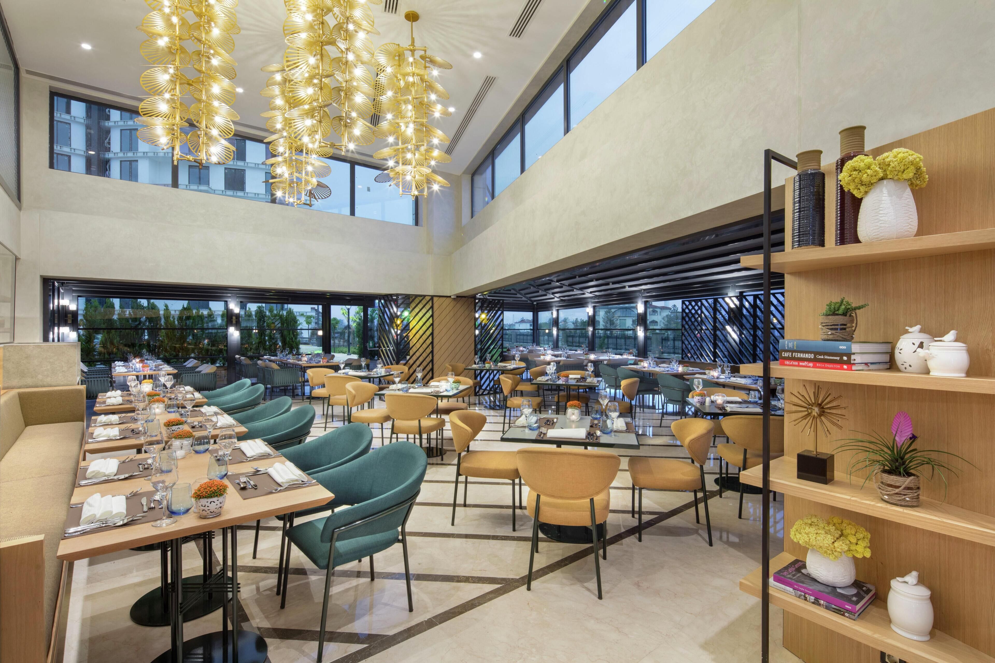 A spacious dining area with multiple tables and chairs, a high ceiling with hanging lights, and large windows providing natural light.