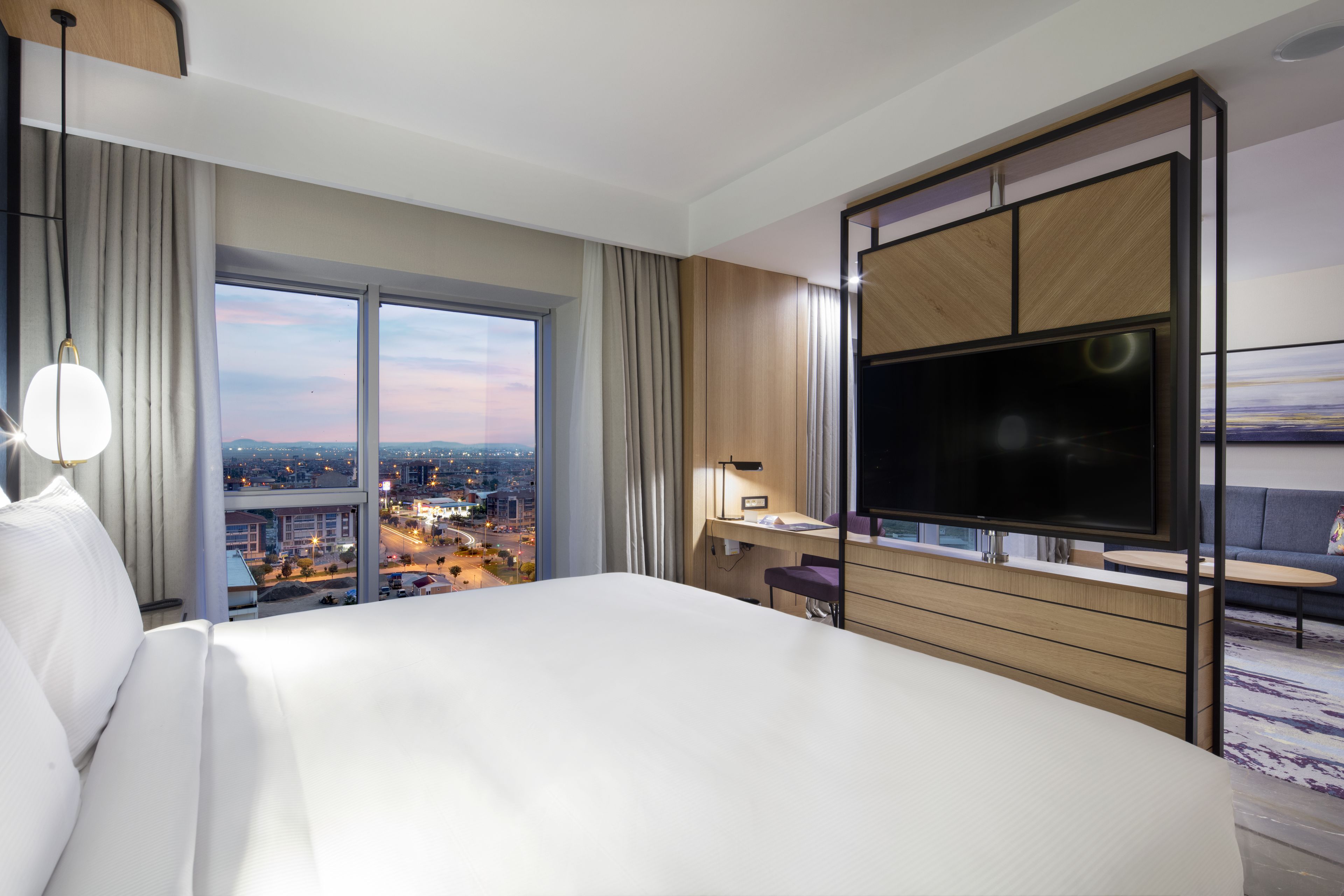 Habitación de hotel con una cama grande, un escritorio, un televisor y vistas a la ciudad al atardecer.