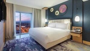 Habitación de hotel con una cama grande, televisión, mesita de noche con teléfono y vista a la ciudad a través de amplios ventanales.